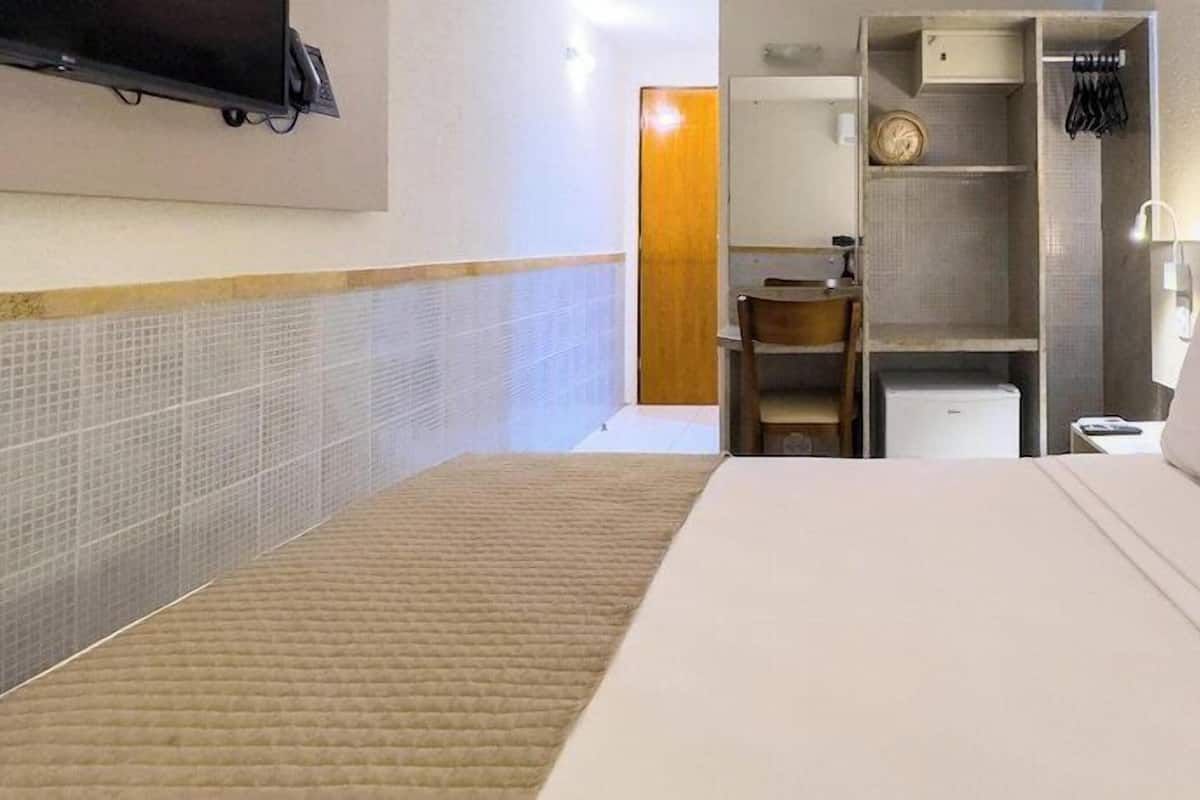 A hotel room with a bed, a TV mounted on the wall, a wardrobe, and a small table with a chair.