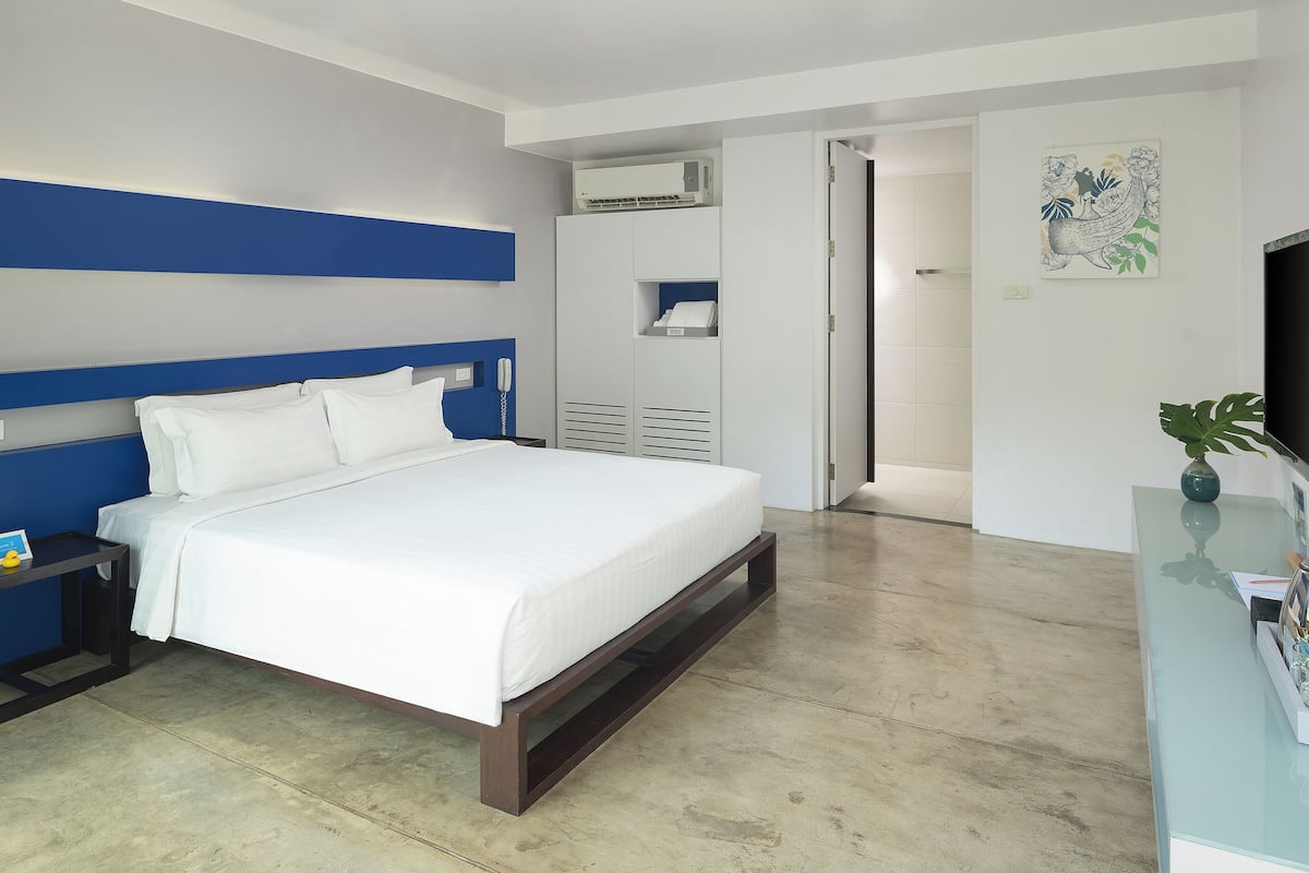 A modern hotel room with a large bed, a bedside table with a phone, a wall-mounted air conditioner, and a flat-screen TV on a glass table.
