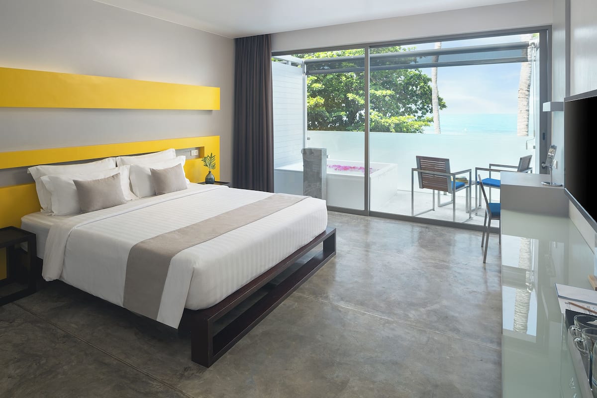 A modern hotel room with a large bed, a nightstand, a desk with a chair, and a balcony with a view of the ocean.