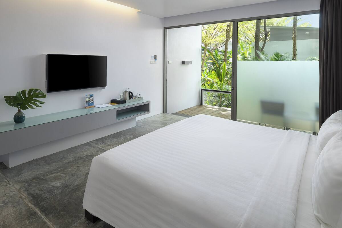 A modern hotel room with a large bed, a flat-screen TV mounted on the wall, a small desk with a plant, and a sliding glass door leading to a balcony with greenery.