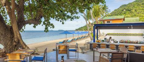 A beachside restaurant with outdoor seating, a bar, and a view of the sea.