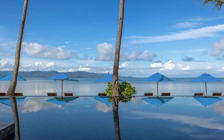 An infinity pool with clear water reflecting the sky and palm trees. There are blue umbrellas and wooden chairs around the pool.