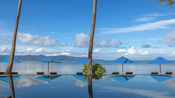 An infinity pool with clear water reflecting the sky and palm trees. There are blue umbrellas and wooden chairs around the pool.