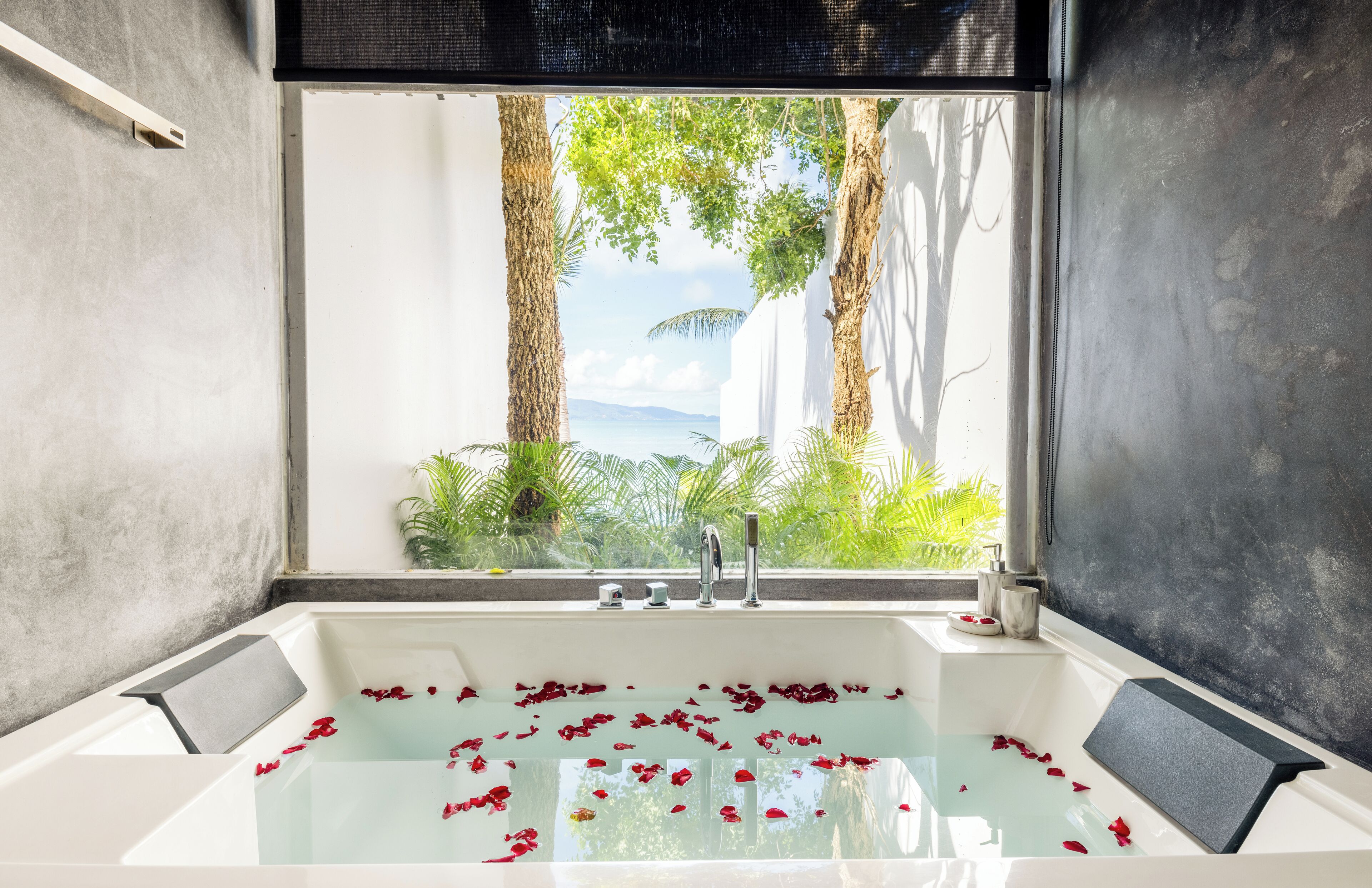 A modern bathroom with a bathtub filled with rose petals, a window with a view of greenery and the sea, and a minimalist design.