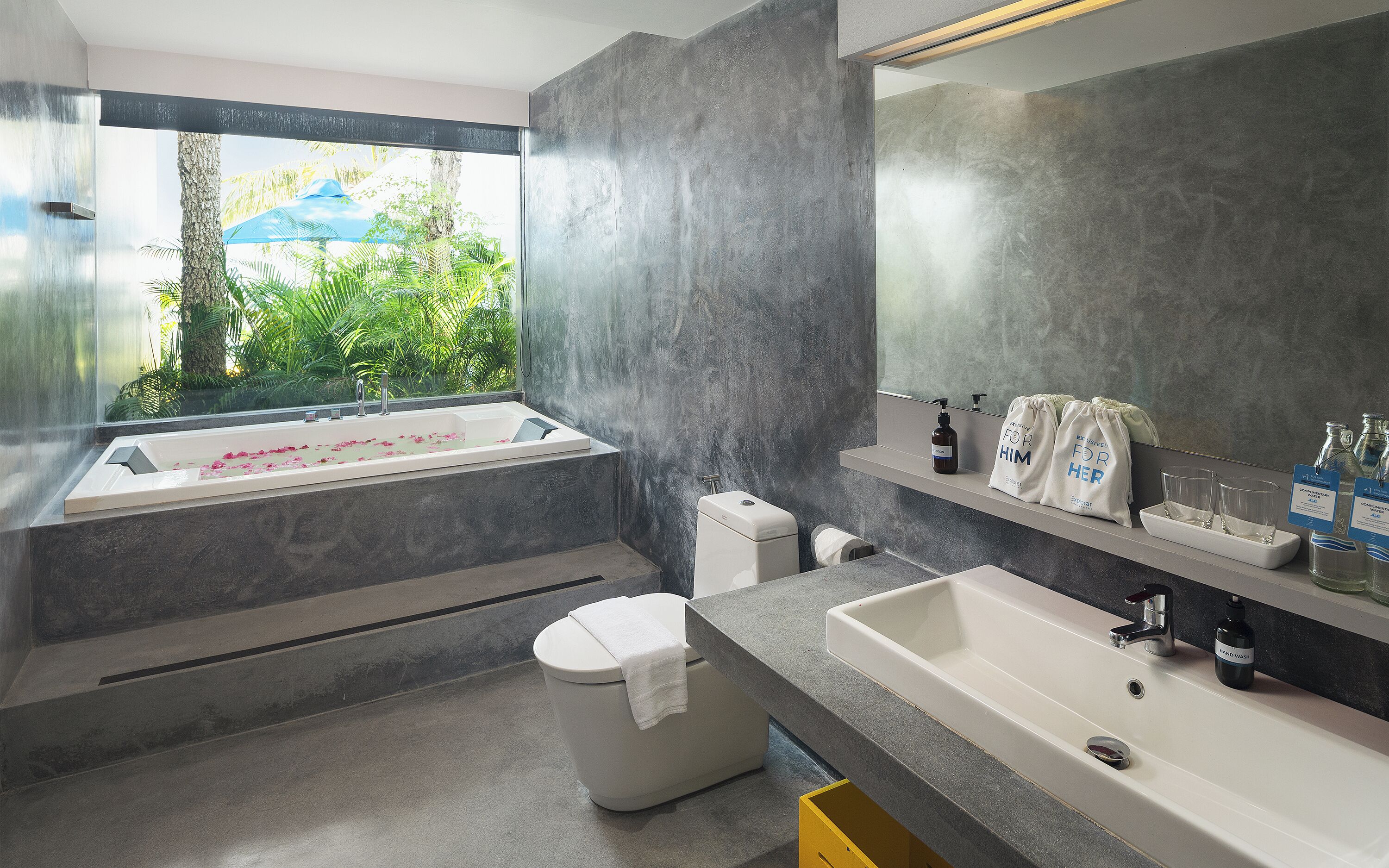 A modern bathroom with a bathtub, a sink, and a toilet. There is a window with a view of greenery outside.
