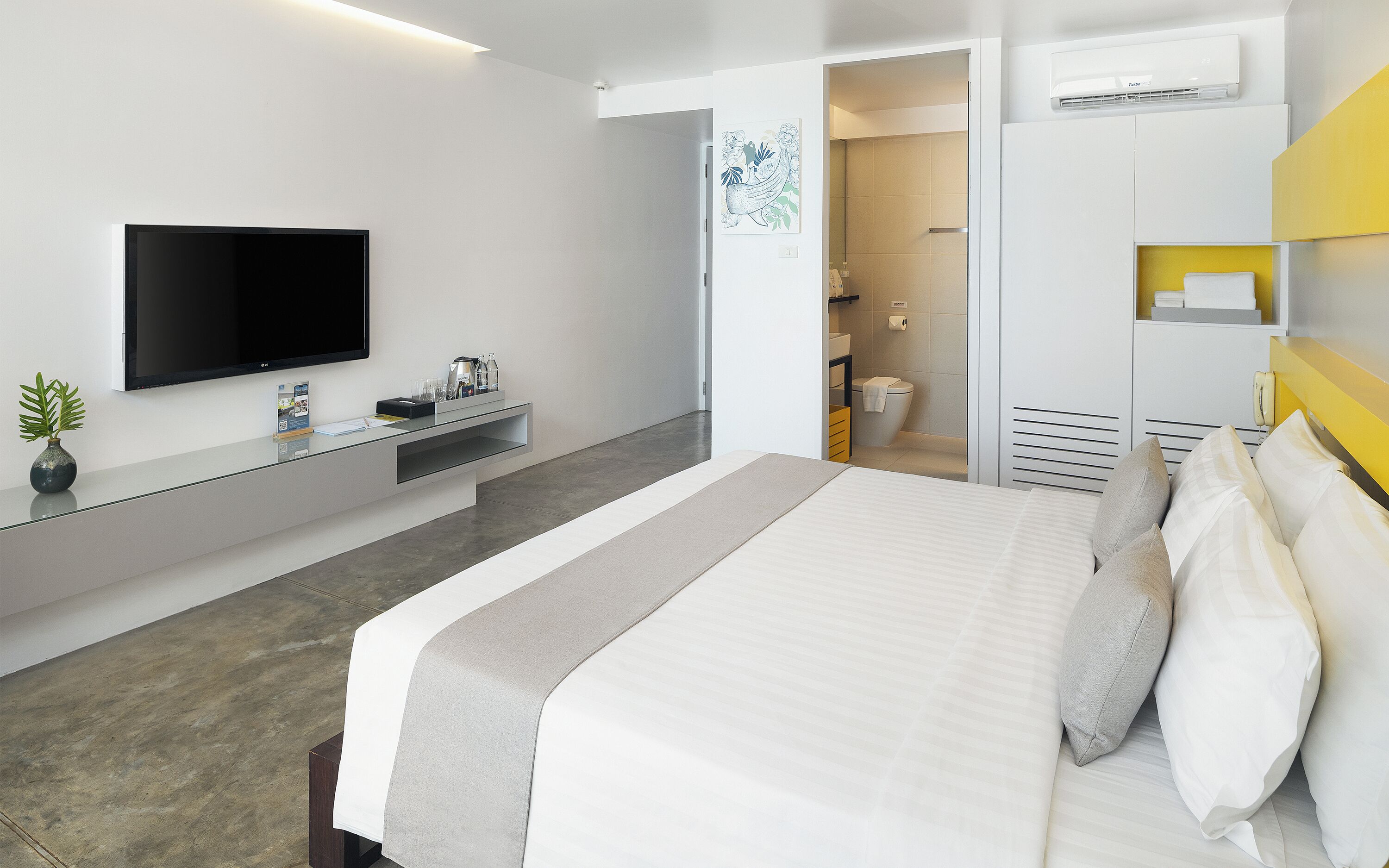 A modern hotel room with a bed, a flat-screen TV, a desk with a vase, and a bathroom visible through an open door.