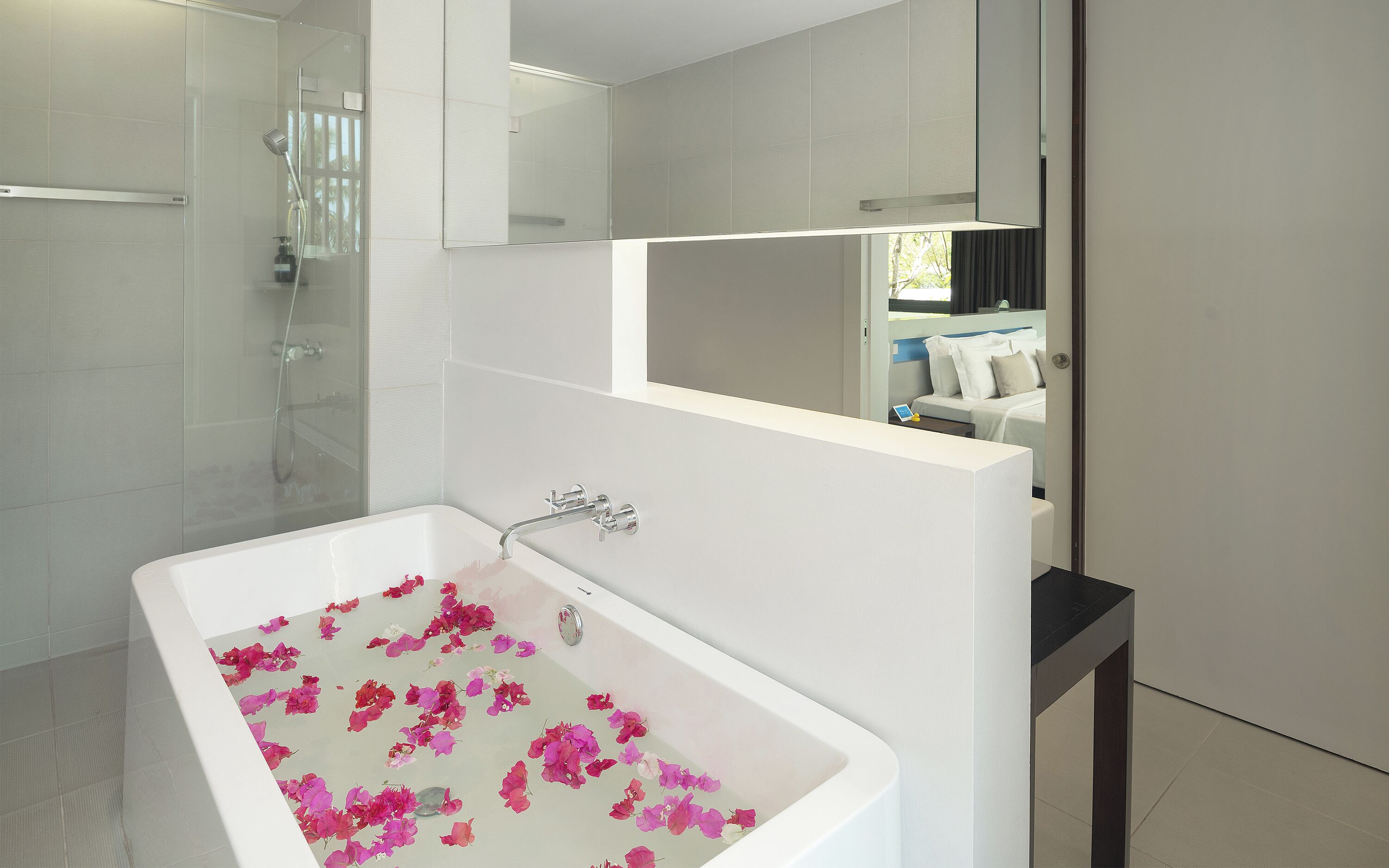 A modern bathroom with a large bathtub filled with rose petals, a glass shower enclosure, and a mirror reflecting a bedroom area.
