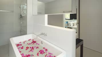 Une salle de bain moderne avec une grande baignoire remplie de pétales de roses, une douche en verre et un miroir qui reflète une partie de la chambre à coucher.