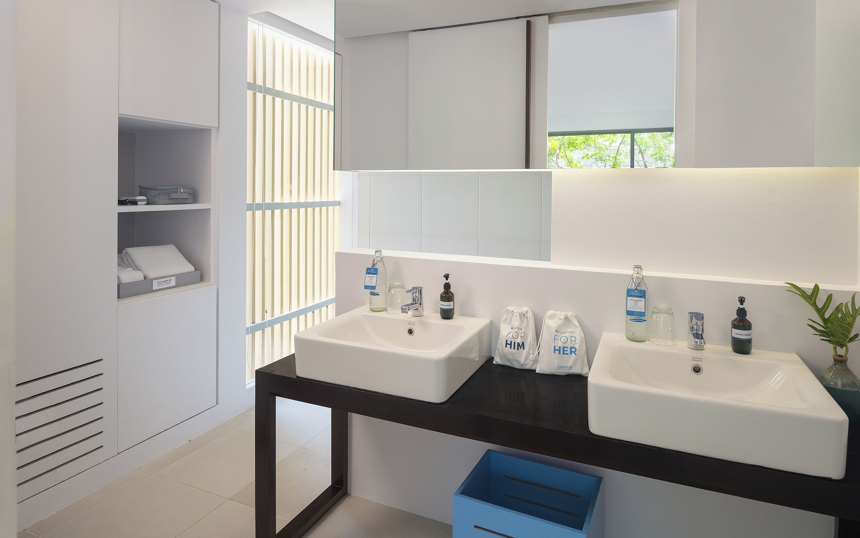 A modern bathroom with two sinks, a mirror, and a window with blinds. There are toiletries on the counter and a blue storage box on the floor.