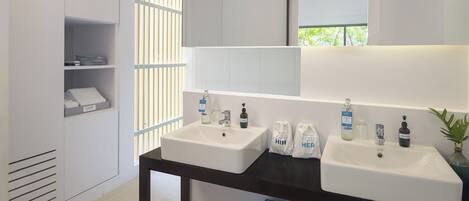 A modern bathroom with two sinks, a mirror, and a window with blinds. There are toiletries on the counter and a blue storage box on the floor.