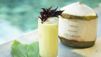 A glass of yellow beverage with a decorative leaf, a whole coconut, and a green leaf on a surface near a body of water.