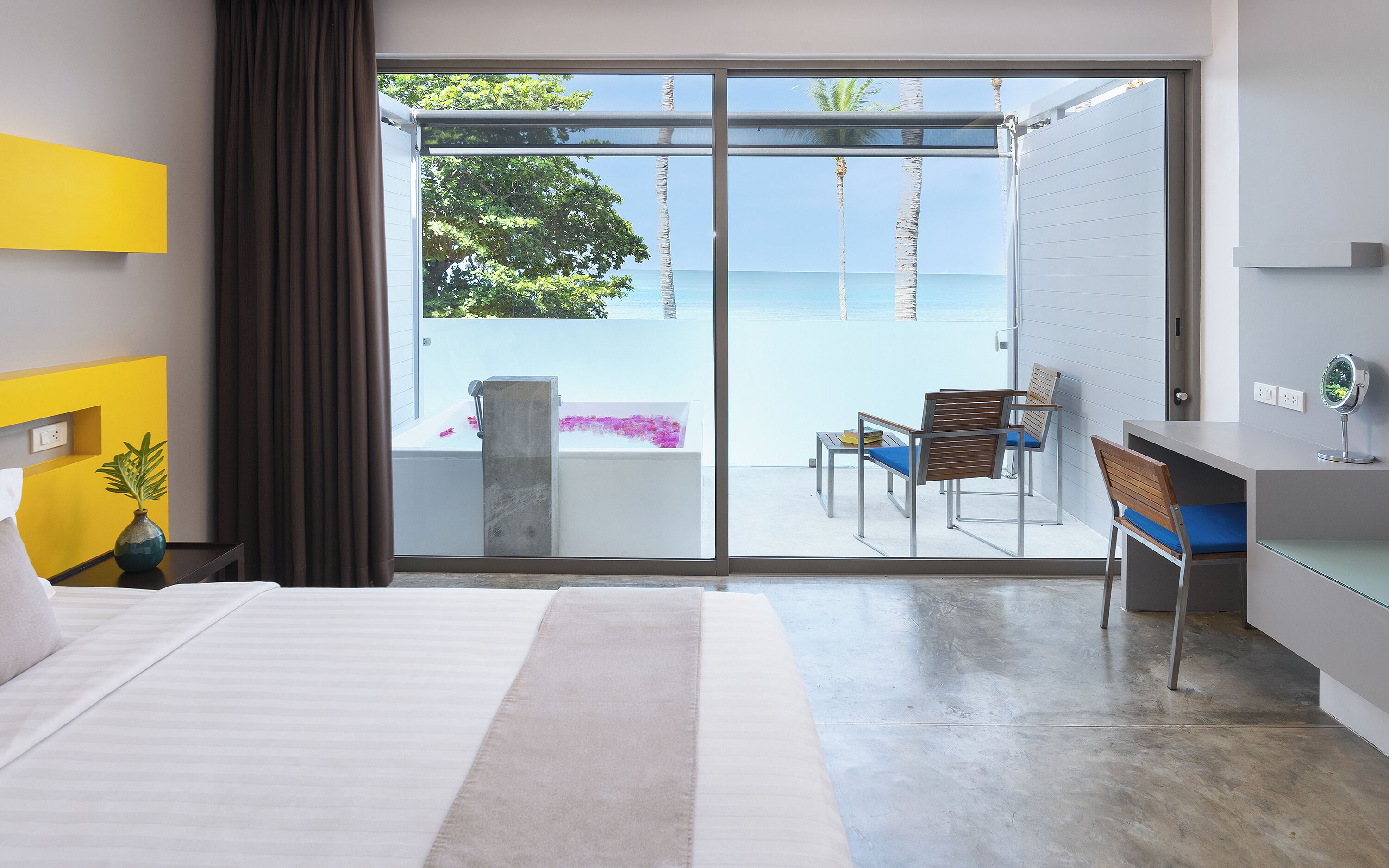 Ein modernes Hotelzimmer mit einer großen Schiebeglasdoor, die auf einen Balkon mit Blick auf das Meer und Palmen führt.