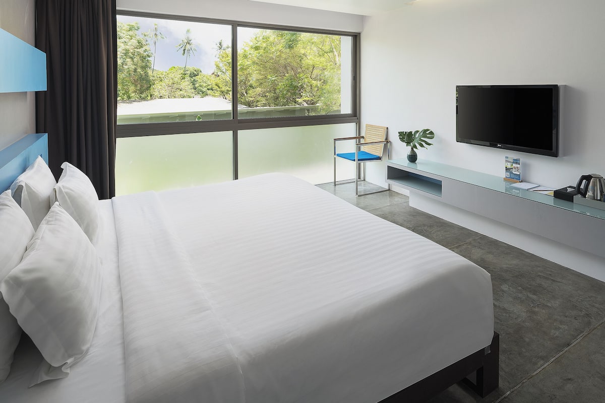 A hotel room with a bed, a TV, a desk with a plant, and a window with a view of trees.