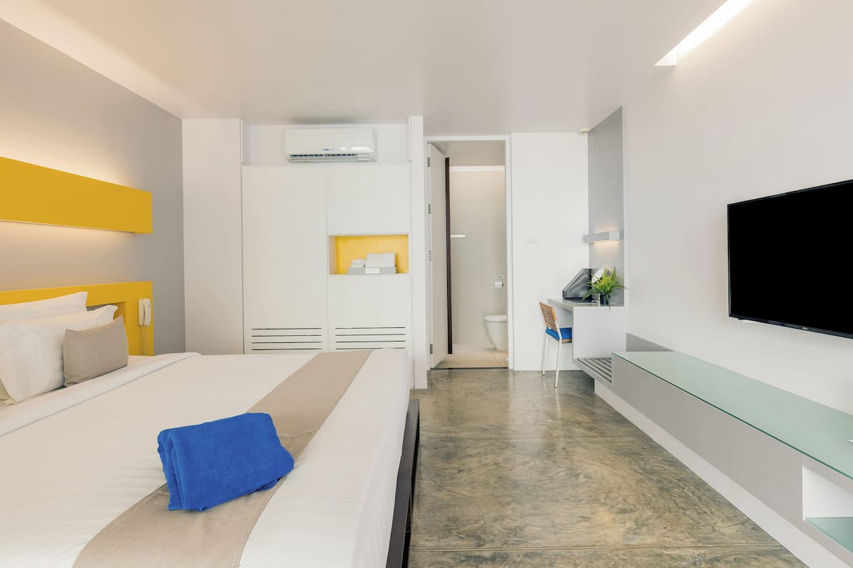 A modern hotel room with a bed, a flat-screen TV, a desk with a chair, and a bathroom visible through an open door.