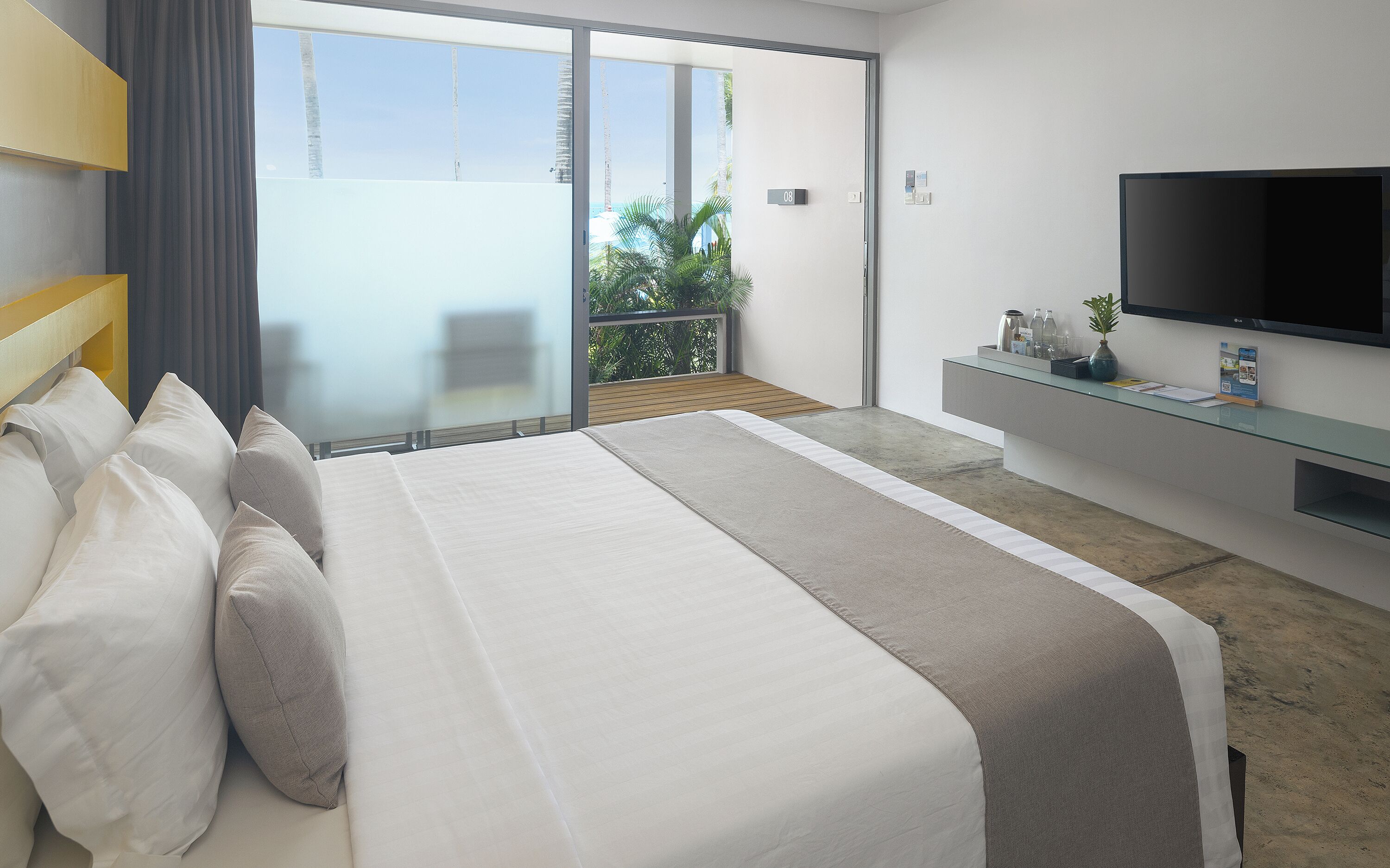 A modern hotel room with a large bed, a flat-screen TV, a glass door leading to a balcony, and a small desk with a vase and a plant.