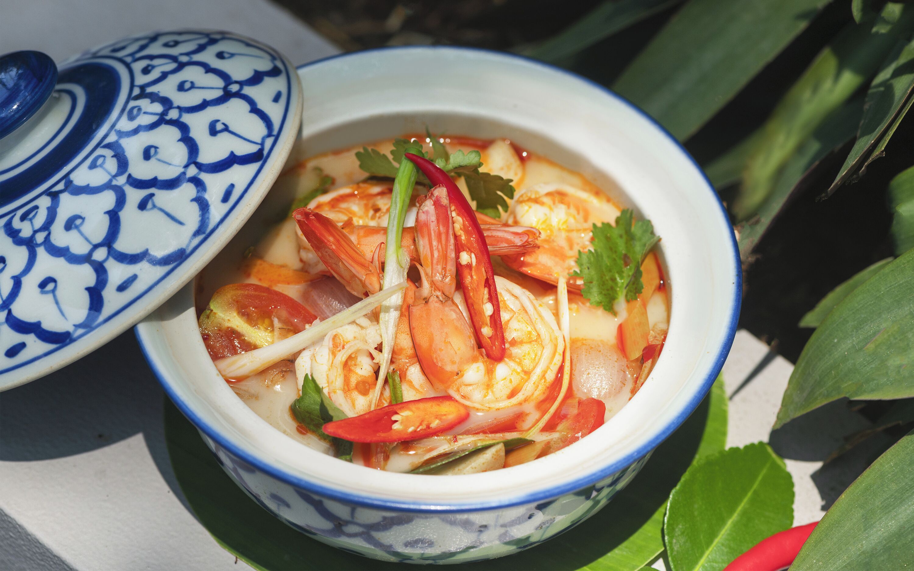 Una zuppa con gamberi, verdure e erbe aromatiche, servita in una ciotola decorata con motivi blu e bianchi.