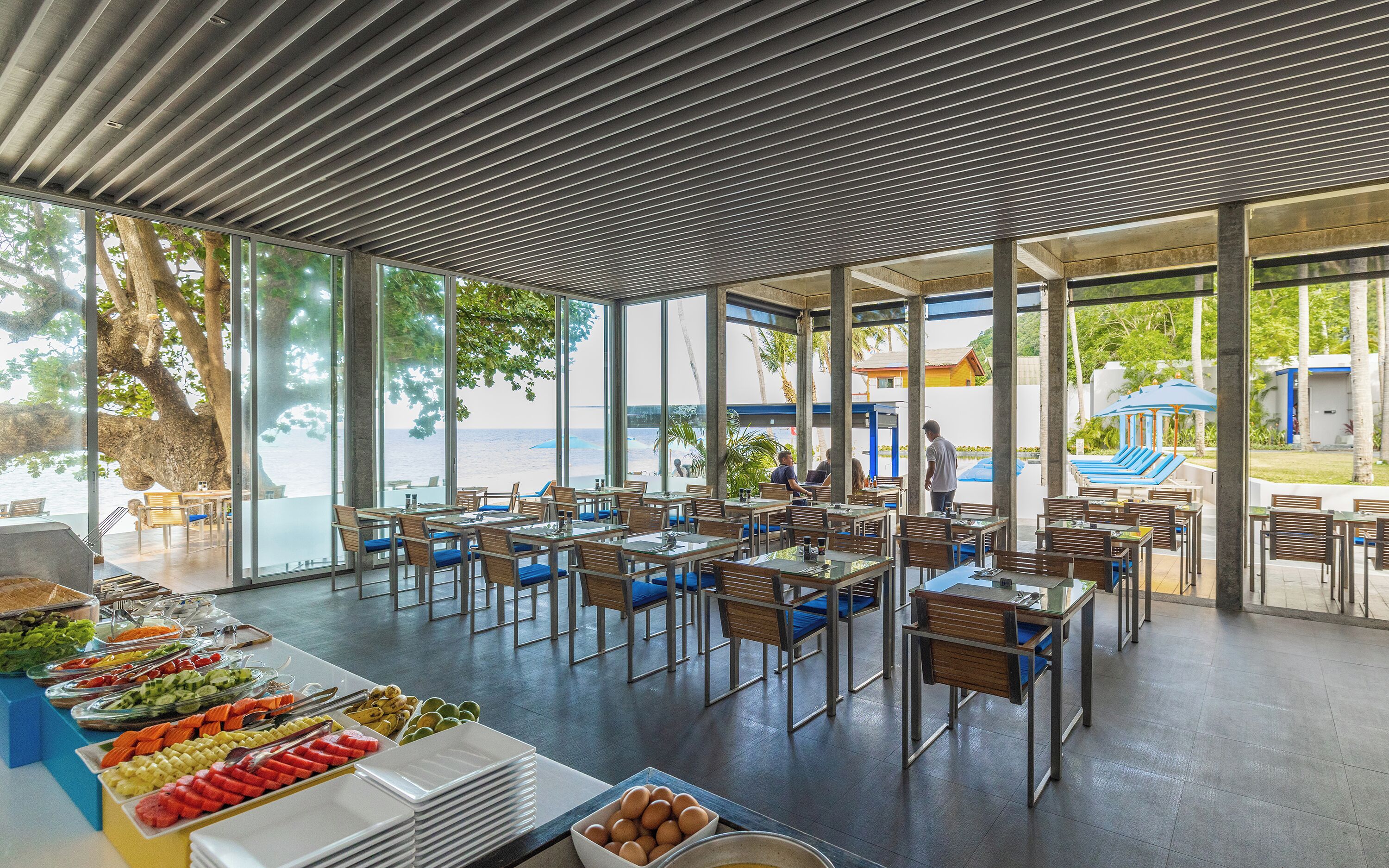 A spacious outdoor dining area with tables and chairs, large windows offering views of the beach and ocean, and a buffet with various food items.