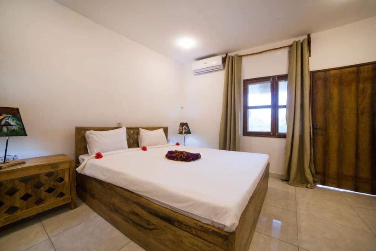 A bedroom with a wooden bed, white bedding, a nightstand with a lamp, and a window with curtains.