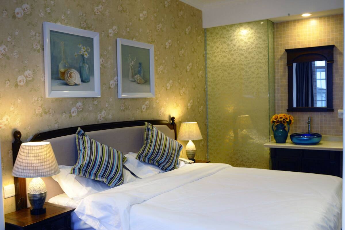 A hotel room with a bed, two bedside lamps, a nightstand, a framed floral artwork, a bathroom with a sink and mirror, and a window with curtains.