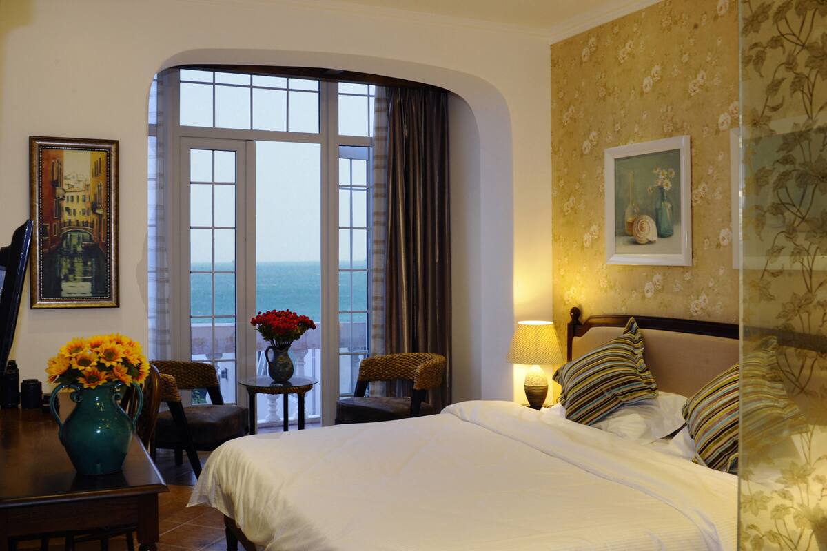 A hotel room with a large window, a bed, a chair, a table with a vase of flowers, and a lamp.