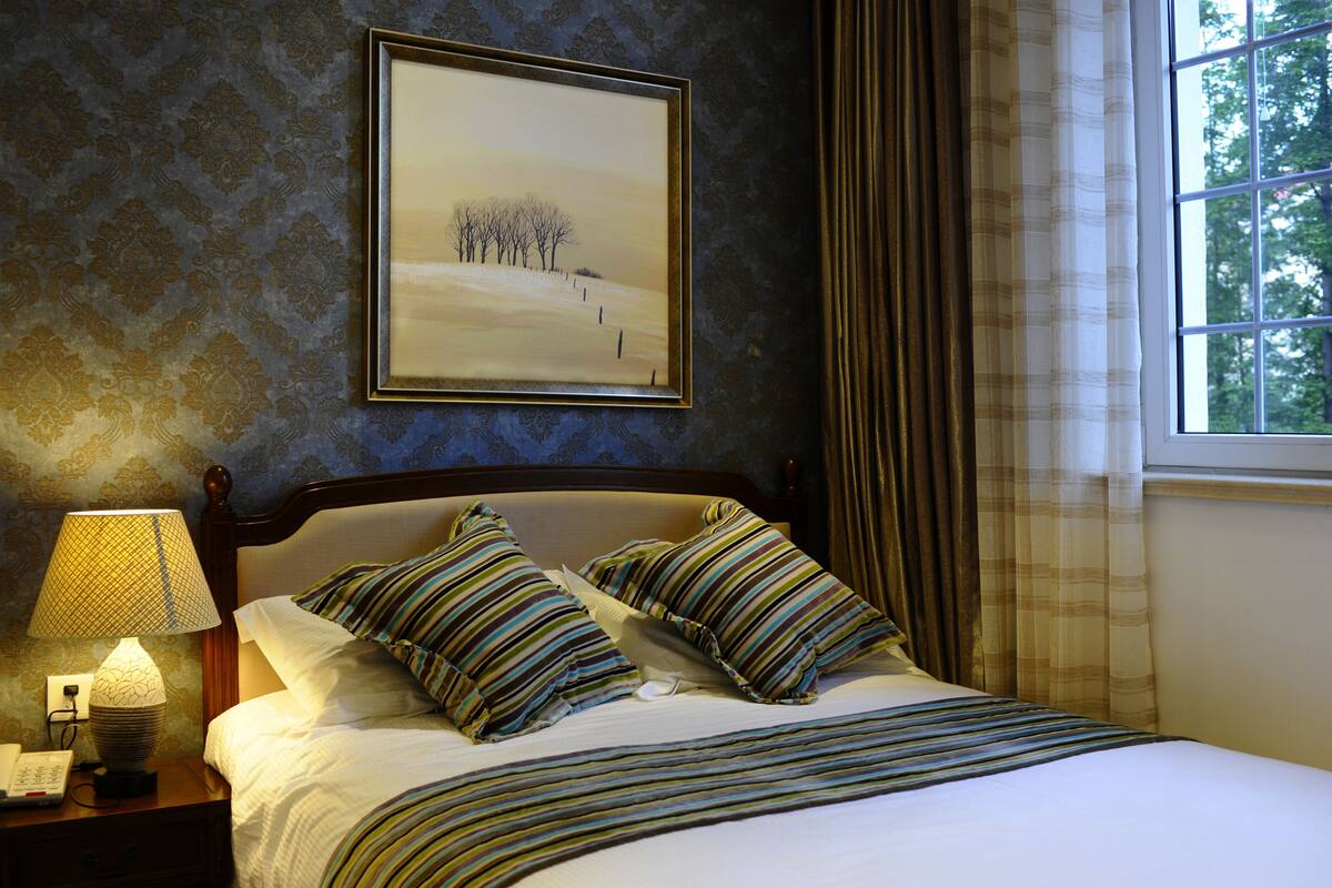 A neatly made bed with striped pillows, a framed landscape painting on the wall, and a window with curtains.