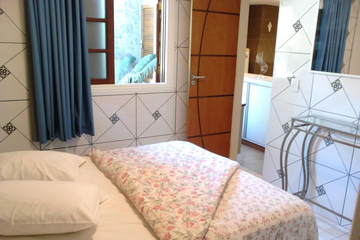 Ein Schlafzimmer mit Bett, einem Fenster mit Vorhängen, einem Badezimmer mit Spiegel und einer Fliesenwand.