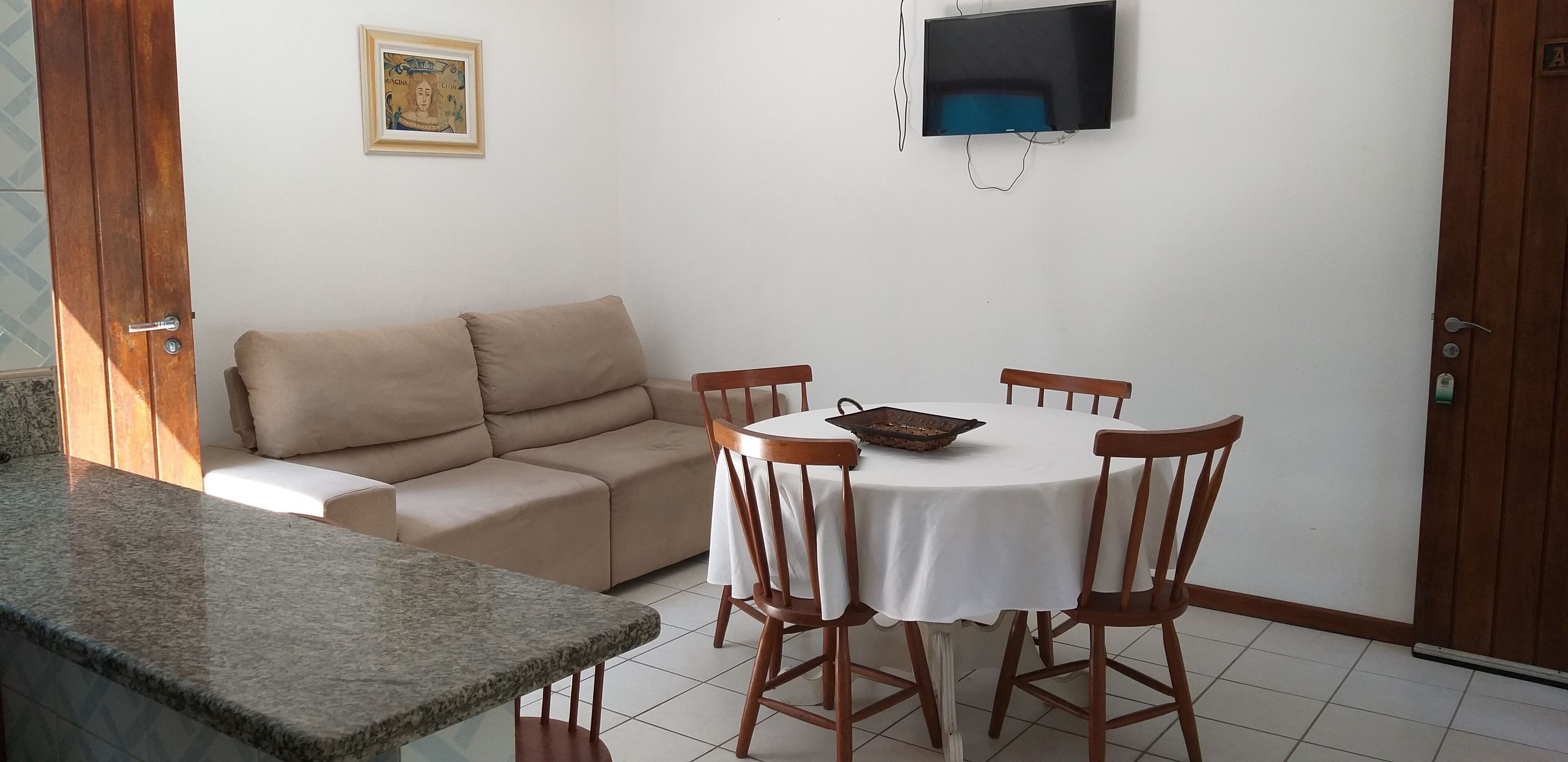 Ein Wohnzimmer mit beiger Couch, einem Esstisch mit vier Stühlen, einem an der Wand montierten Fernseher und einer Granit-Arbeitsplatte.