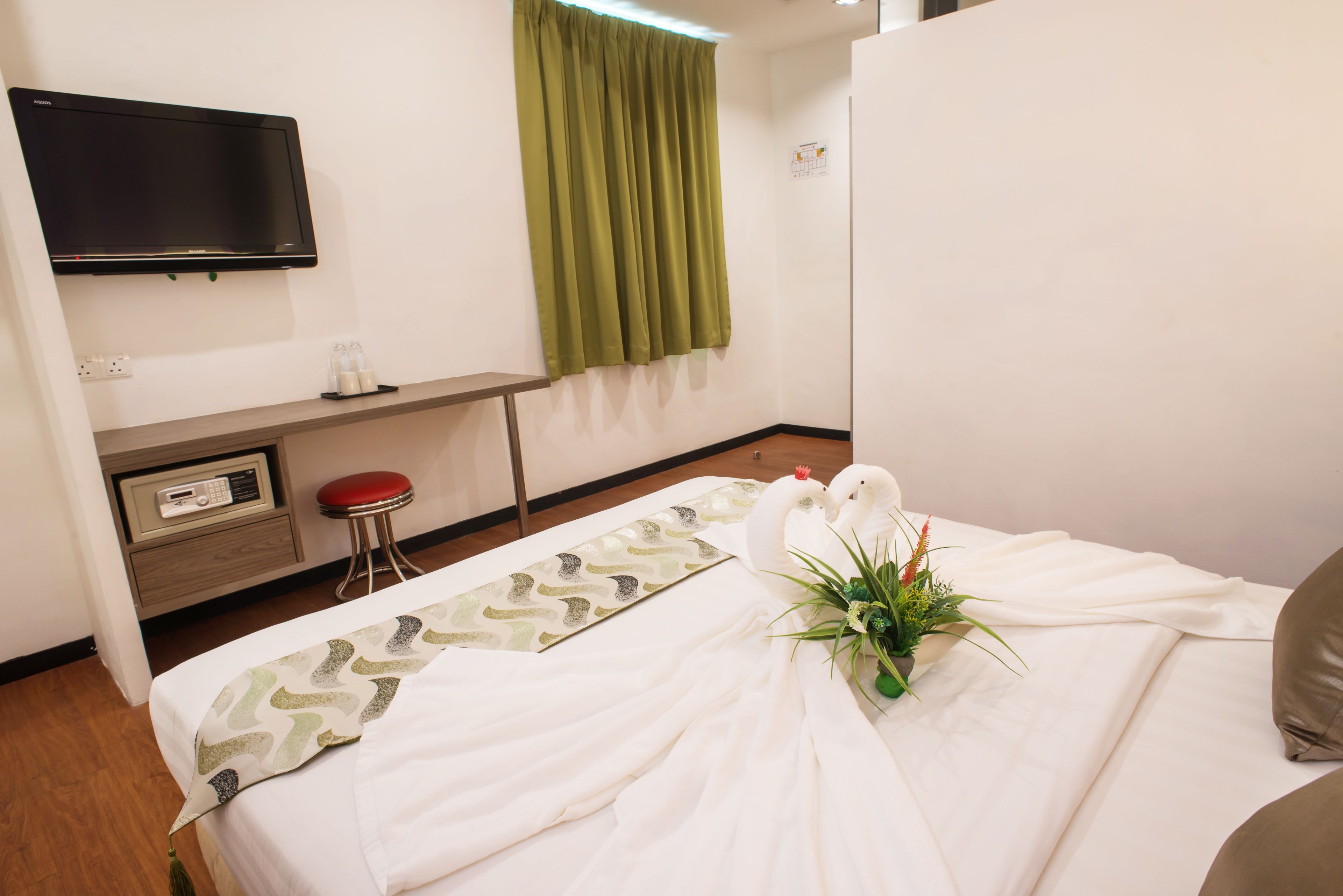 A hotel room with a bed, a desk, a television, a red stool, and a potted plant.