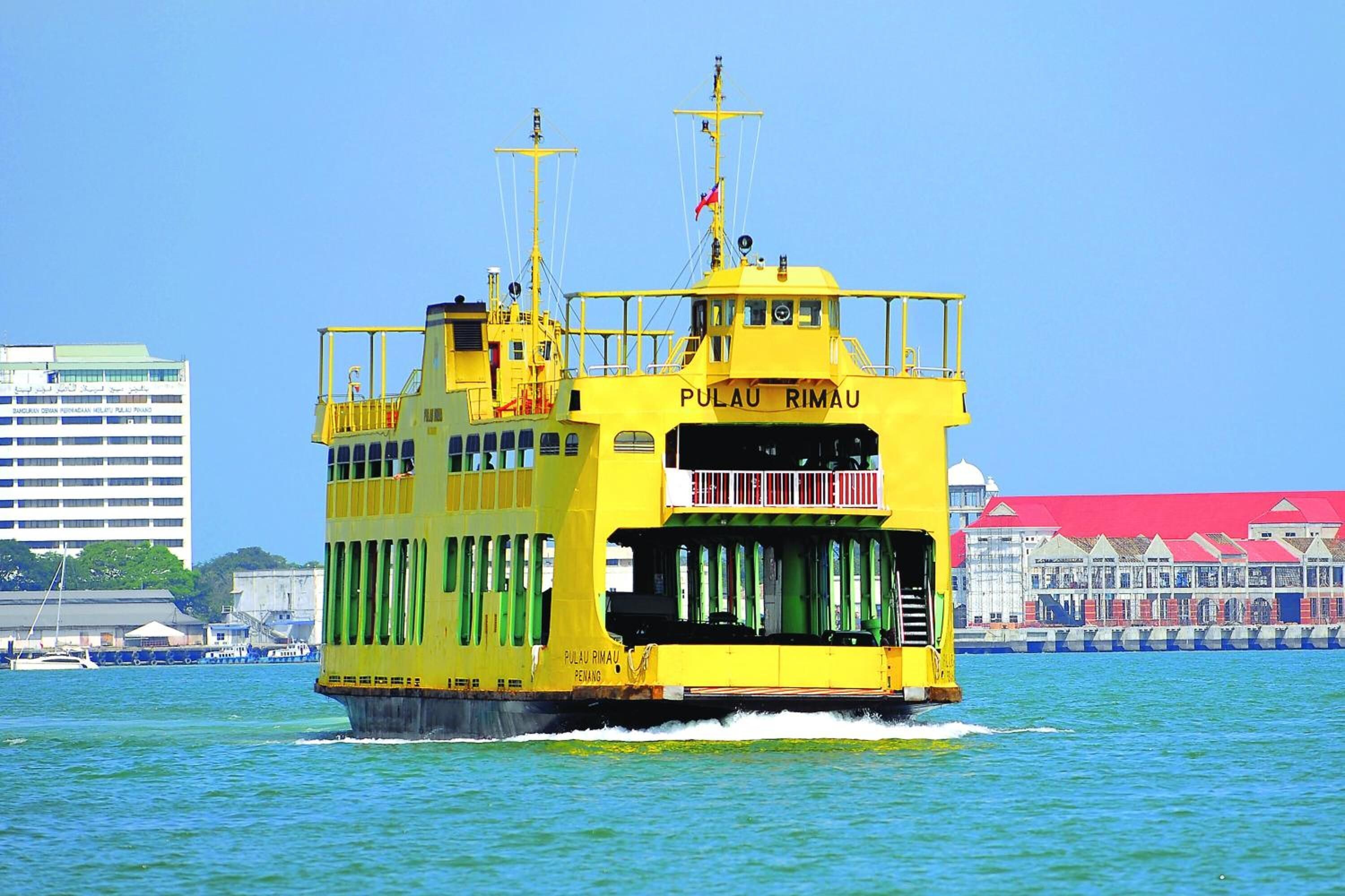 Eine gelbe Fähre namens PULAU RIMAU PENANG mit freier Sicht auf das Wasser und einem Gebäude mit rotem Dach im Hintergrund.