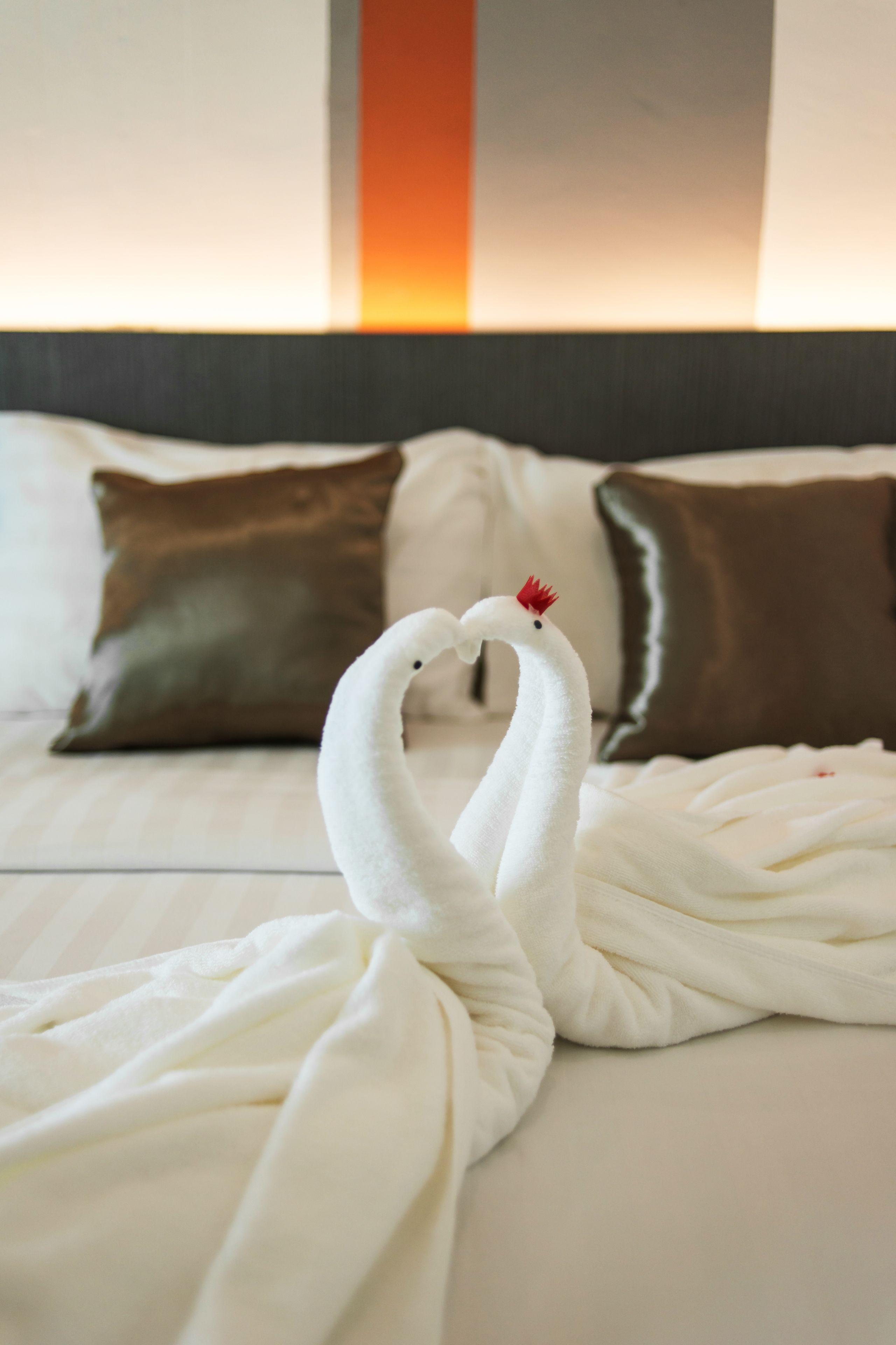 A bed with two white towels arranged to resemble swans, pillows in the background, and a neutral-colored headboard.