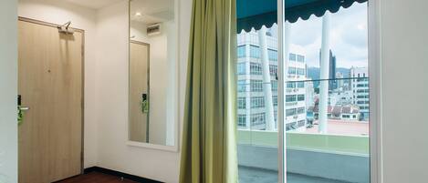 A room with a wooden floor, a sliding glass door with a green curtain, a large mirror, and a view of buildings through the door.