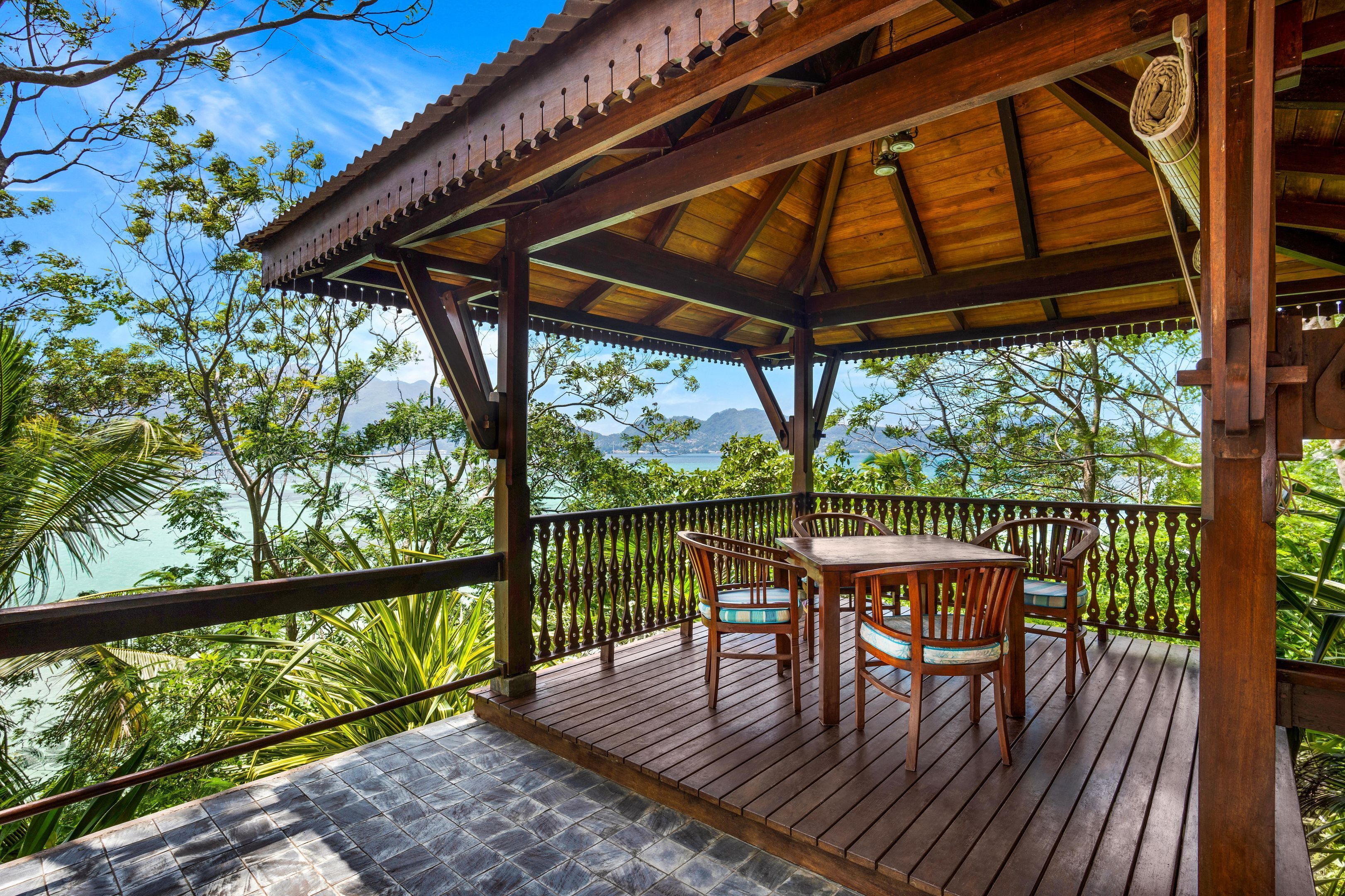 A wooden deck with a table and chairs, overlooking a tropical landscape with trees and a view of the sea.