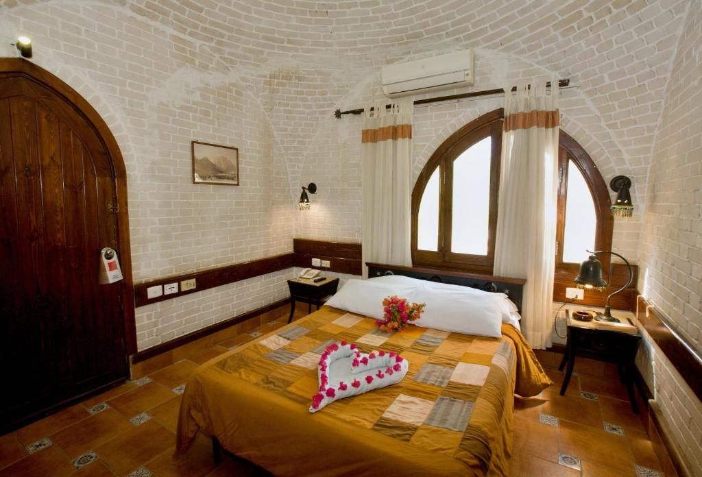 A hotel room with a brick wall, a large wooden door, a bed with a heart-shaped pillow, a nightstand, and a window with curtains.