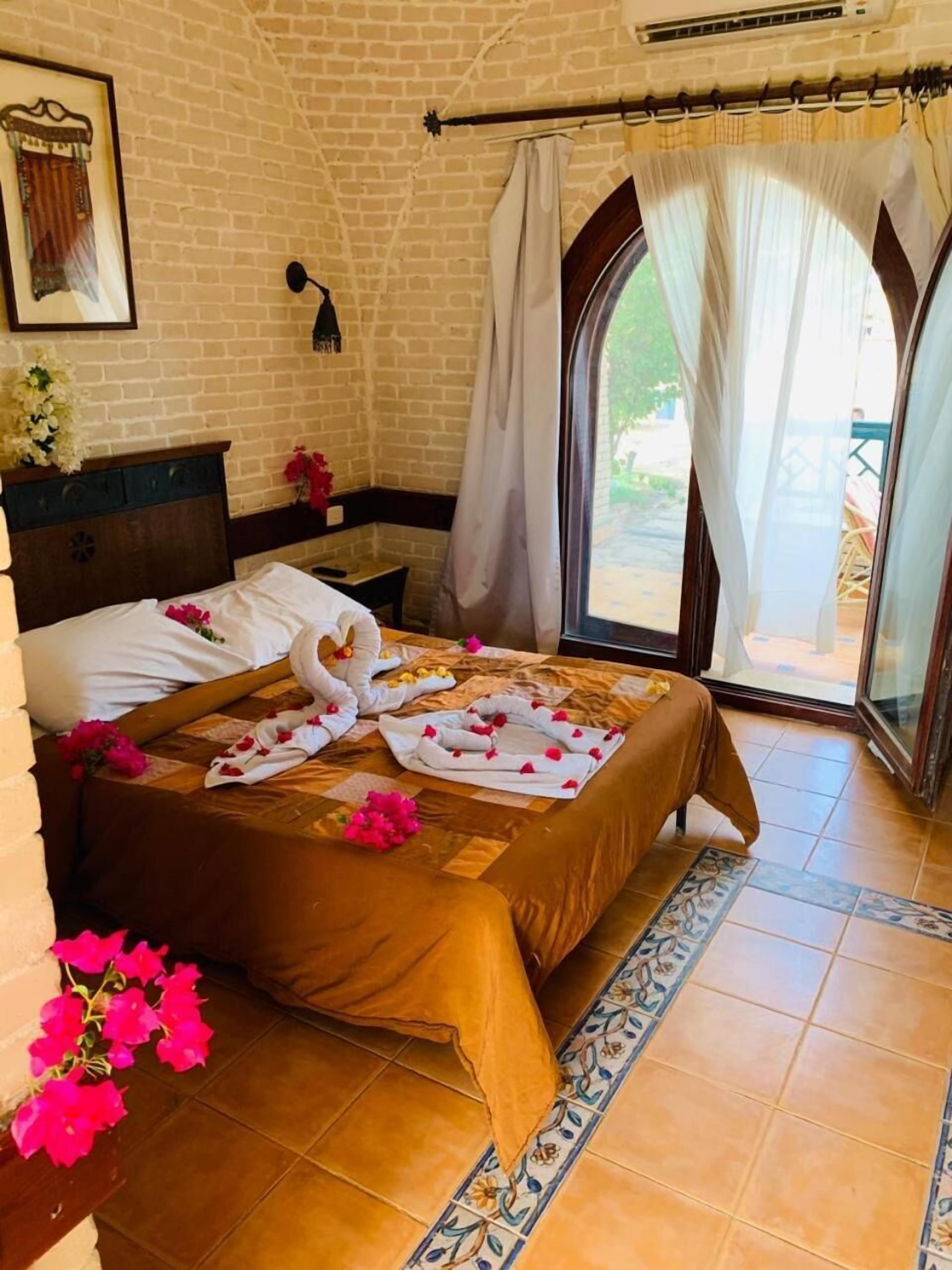 A hotel room with a brick wall, a bed with a brown bedspread and white pillows, a nightstand with a vase of flowers, and a view through a glass door.