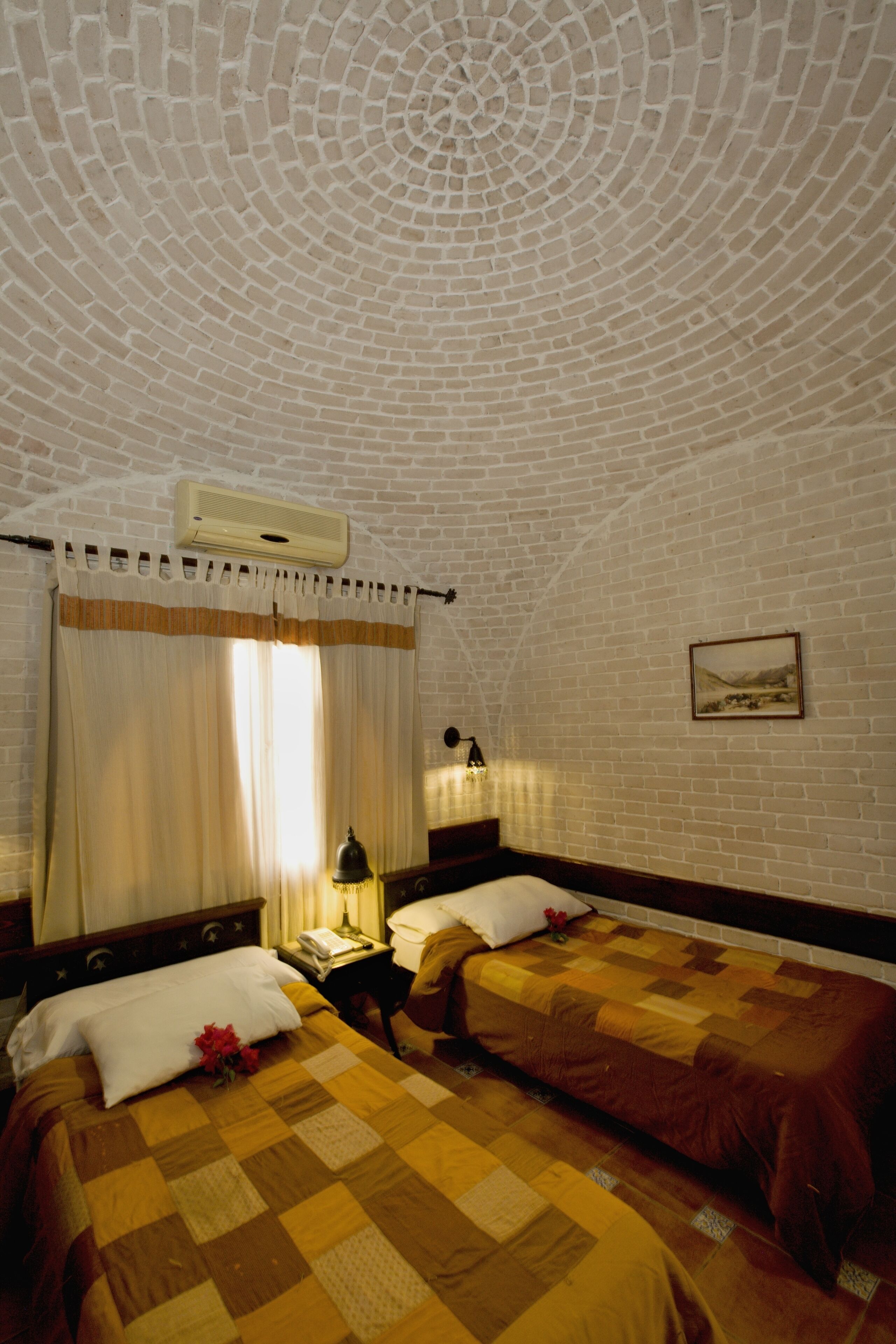 Habitación con dos camas, pared de ladrillo, unidad de aire acondicionado, ventana con cortinas, mesita de noche con lámpara y un cuadro enmarcado en la pared.