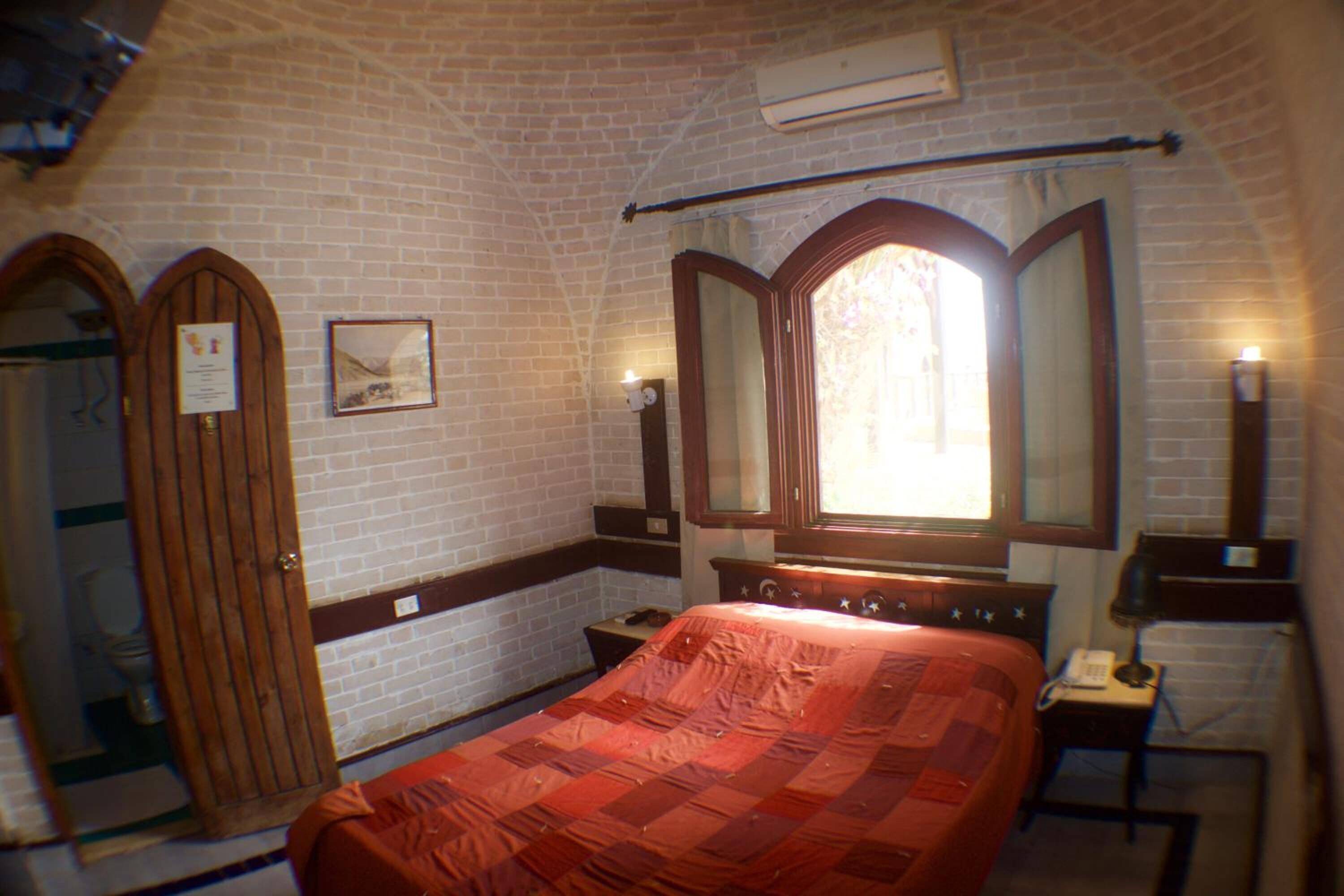 Una habitación con pared de ladrillo, puerta de madera, cama con manta roja, mesita de noche con lámpara y espejo sobre la cama.
