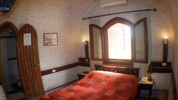 Una habitación con pared de ladrillo, puerta de madera, cama con manta roja, mesita de noche con lámpara y espejo sobre la cama.