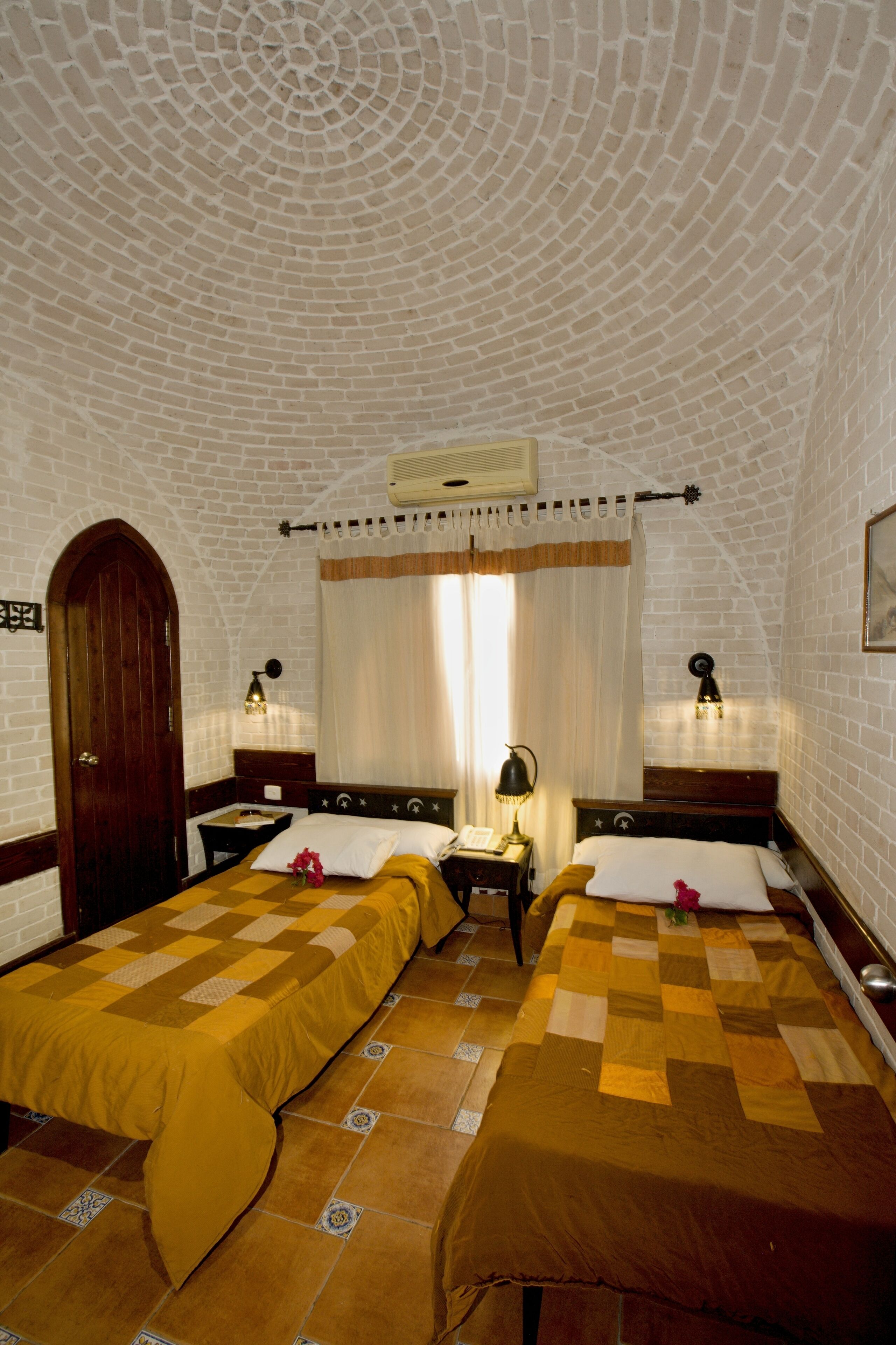 Una habitación con dos camas, una puerta de madera, una ventana con cortinas, un aire acondicionado instalado en la pared y una pequeña mesa con una lámpara.
