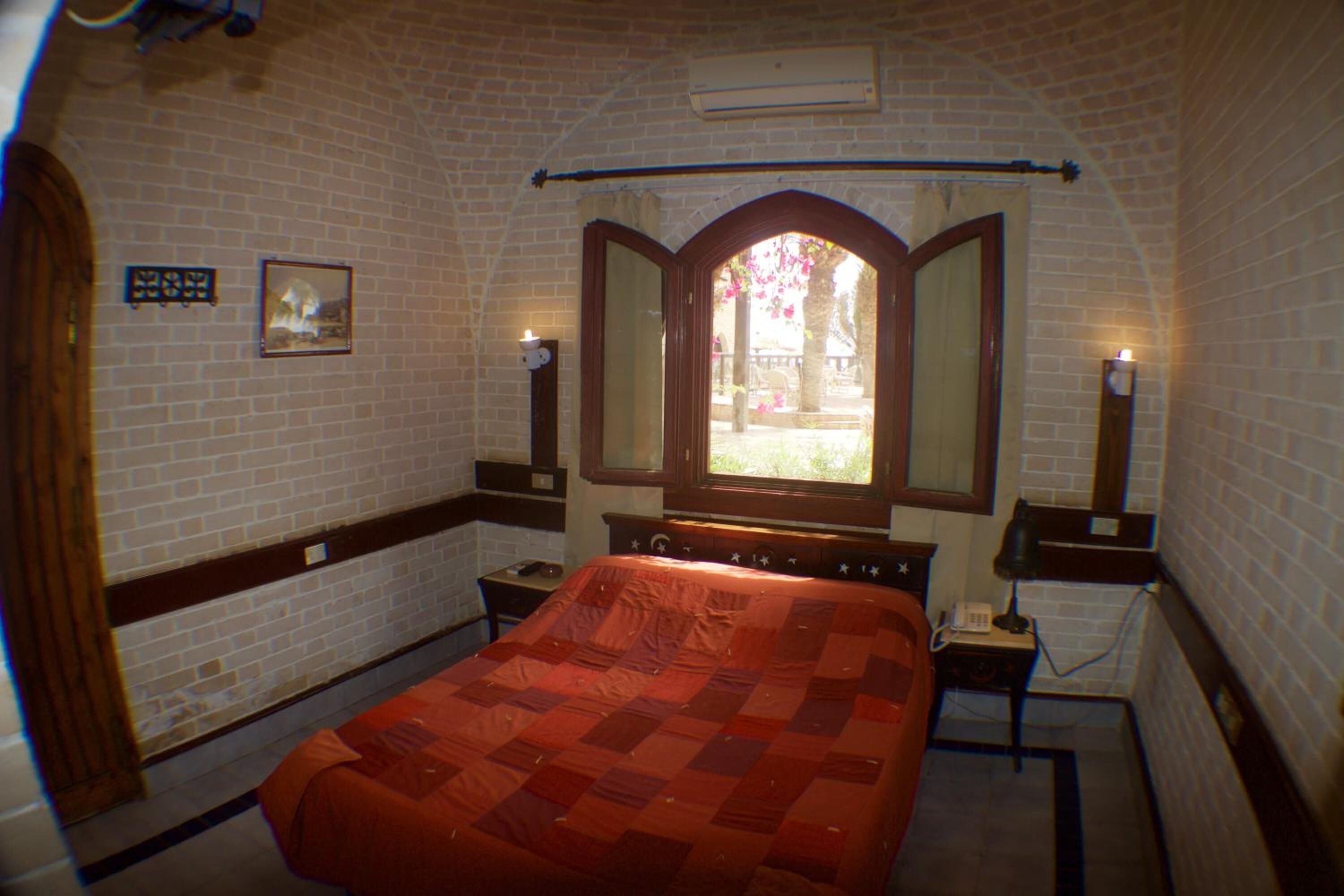 Une chambre avec un mur en briques, un grand lit recouvert d’une couette rouge, deux tables de chevet avec des lampes, une fenêtre à cadre cintré et un tableau accroché au mur.