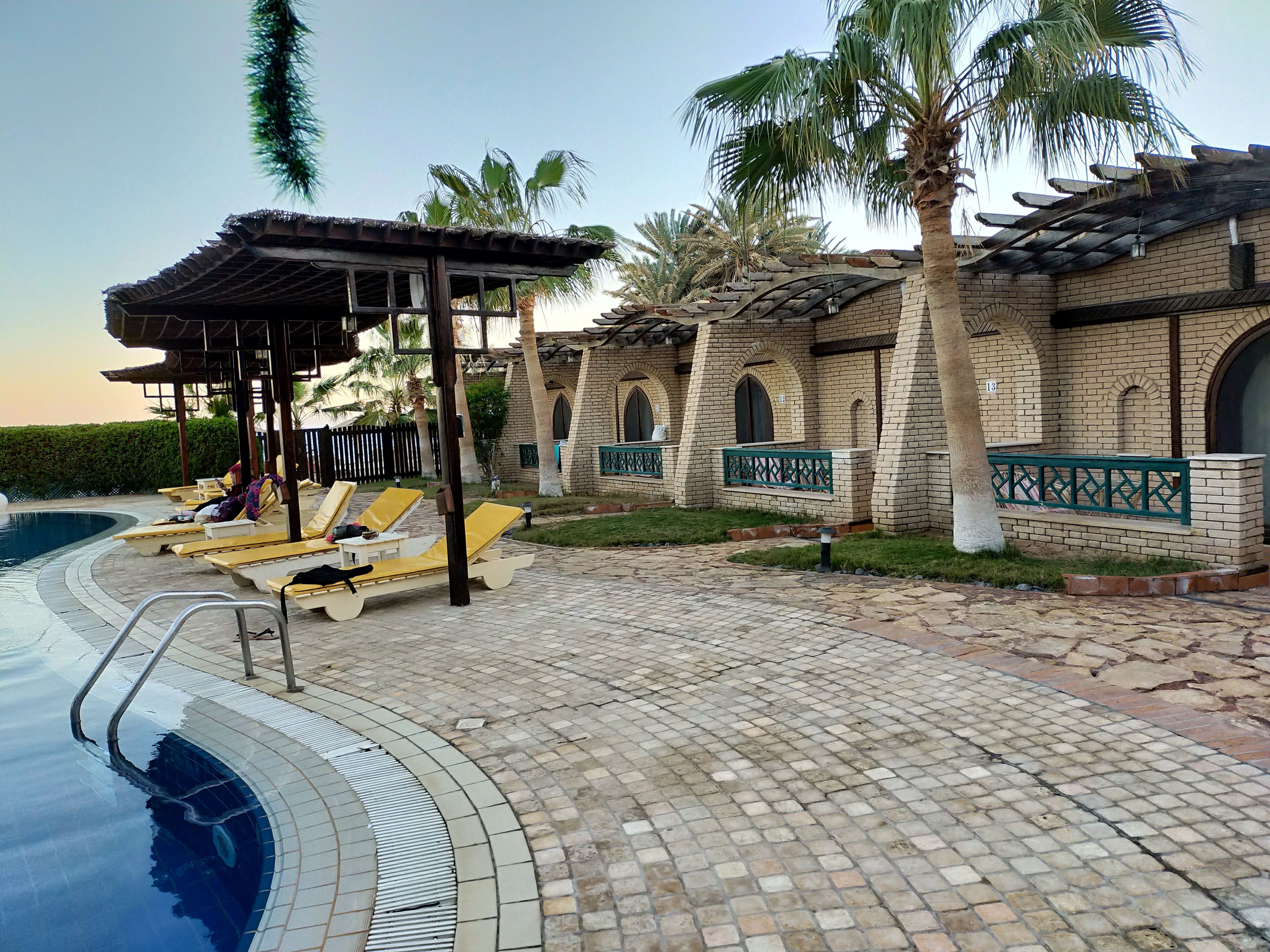 A poolside area with lounge chairs, a pergola, and a brick building with arched windows.