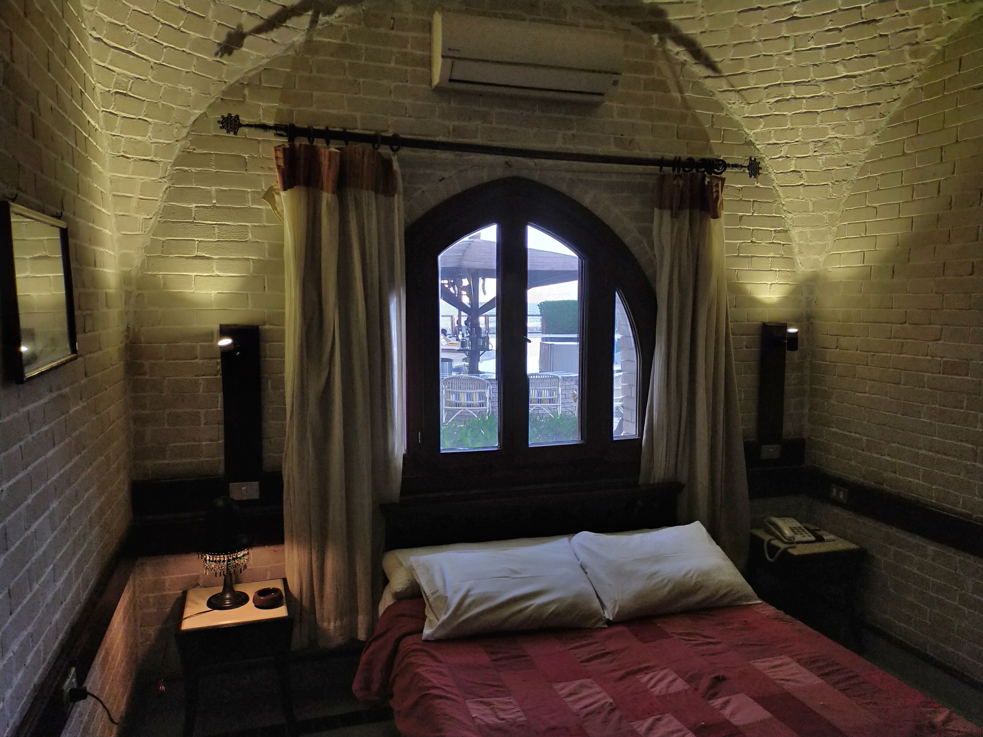 A room with a bed, a window with curtains, a small table with a lamp, and a wall-mounted phone.