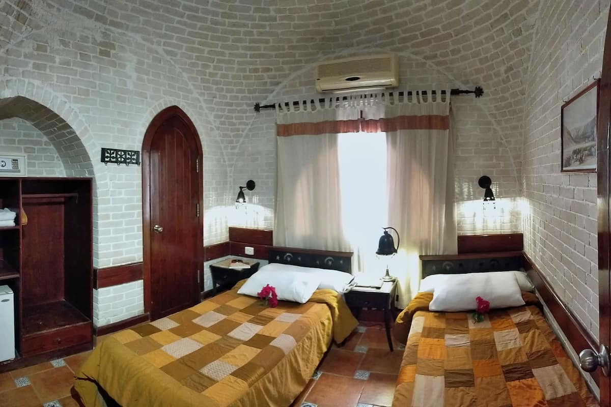 A room with two beds, a wooden wardrobe, a small refrigerator, and a brick wall.