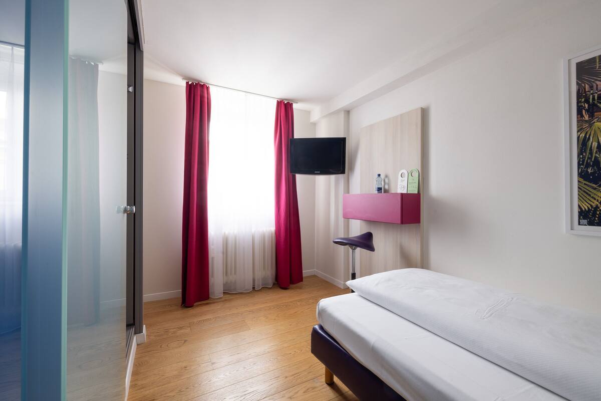 Ein modernes Hotelzimmer mit einem Bett, einem an der Wand montierten Fernseher, einem Nachttisch mit Lampe und einem Fenster mit roten Vorhängen.