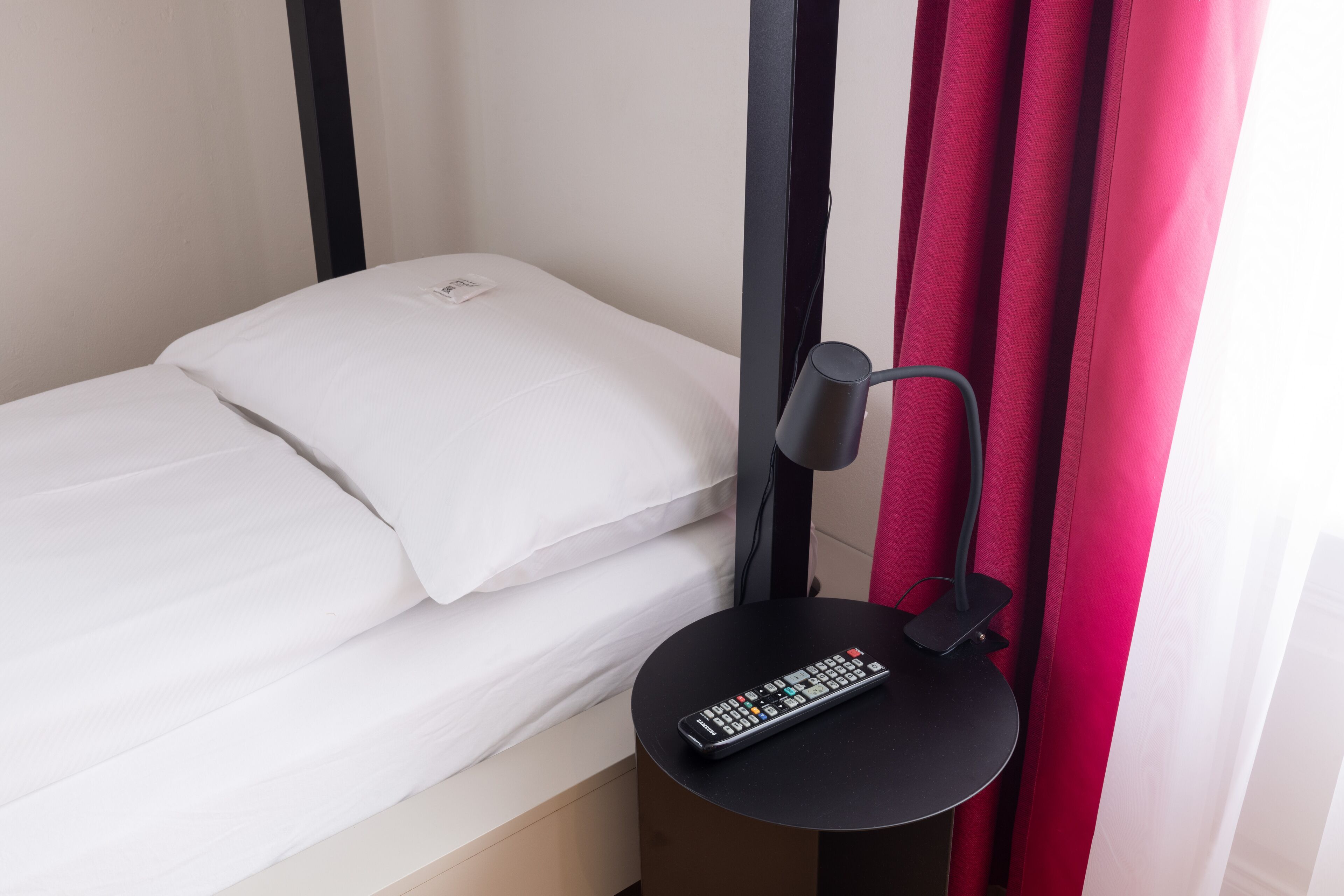 Un lit simple avec une literie blanche, une table de chevet noire avec une télécommande, et un rideau rouge.