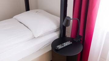 Un lit simple avec une literie blanche, une table de chevet noire avec une télécommande, et un rideau rouge.