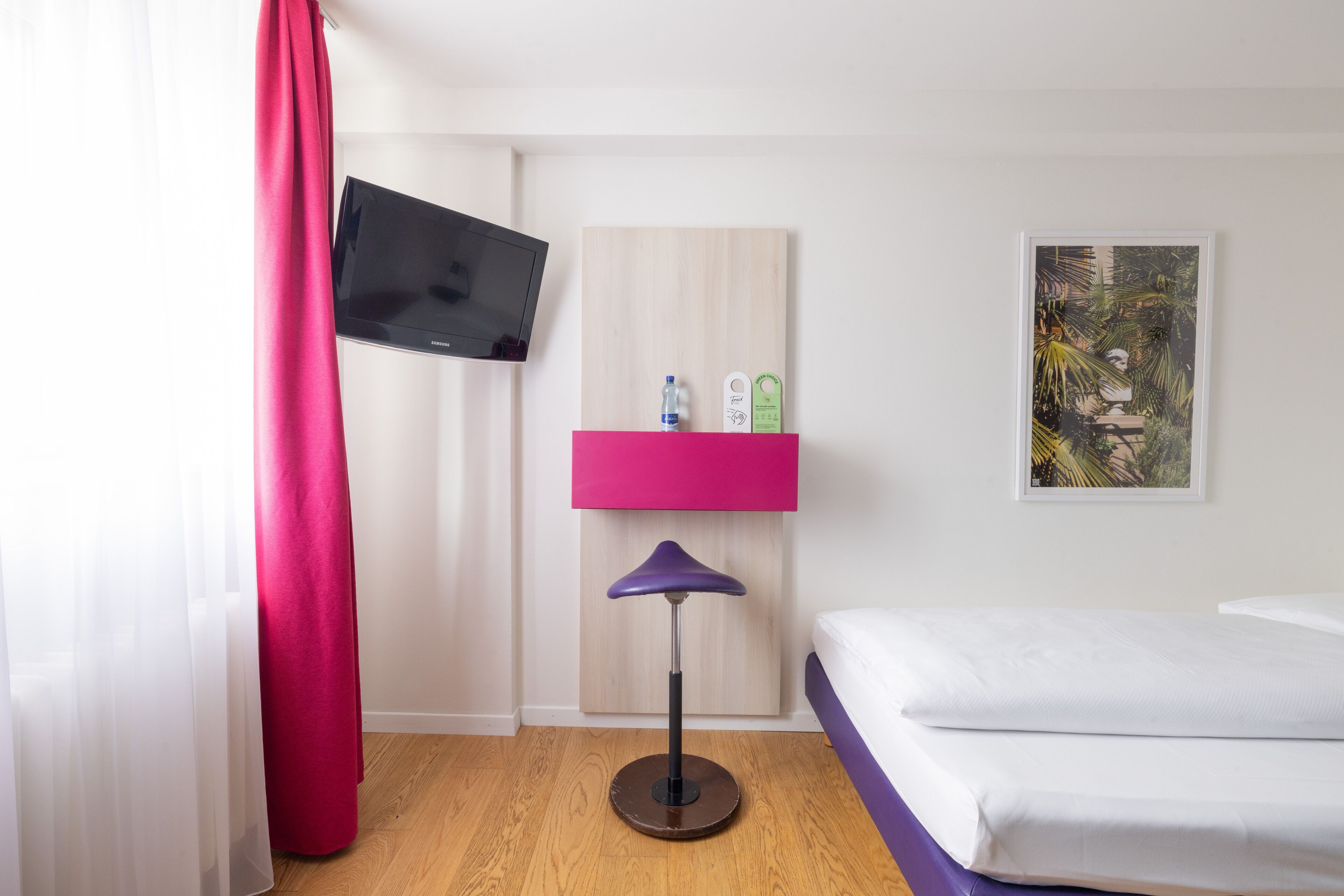 Hotelkamer met een bed, een aan de muur bevestigde televisie, een roze plankje met toiletartikelen en een ingelijst schilderij.