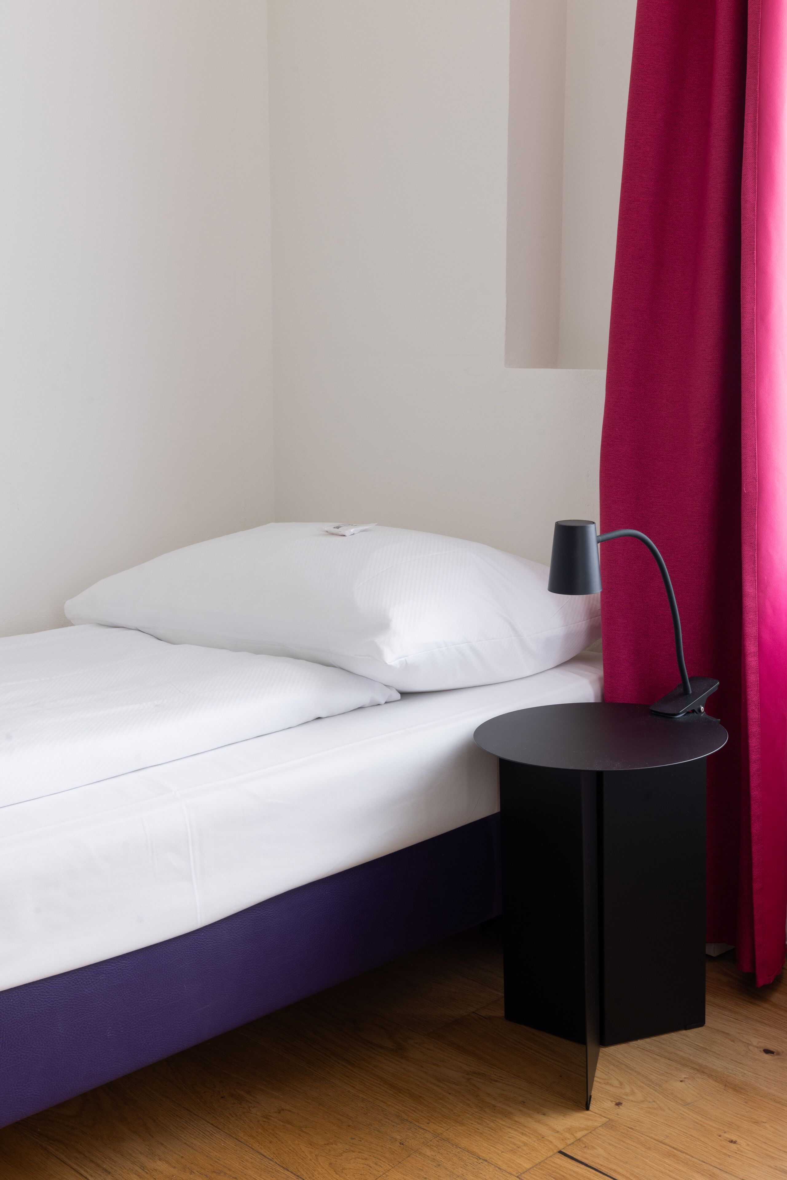 A neatly made bed with white linens, a black side table with a lamp, and a purple bed frame.