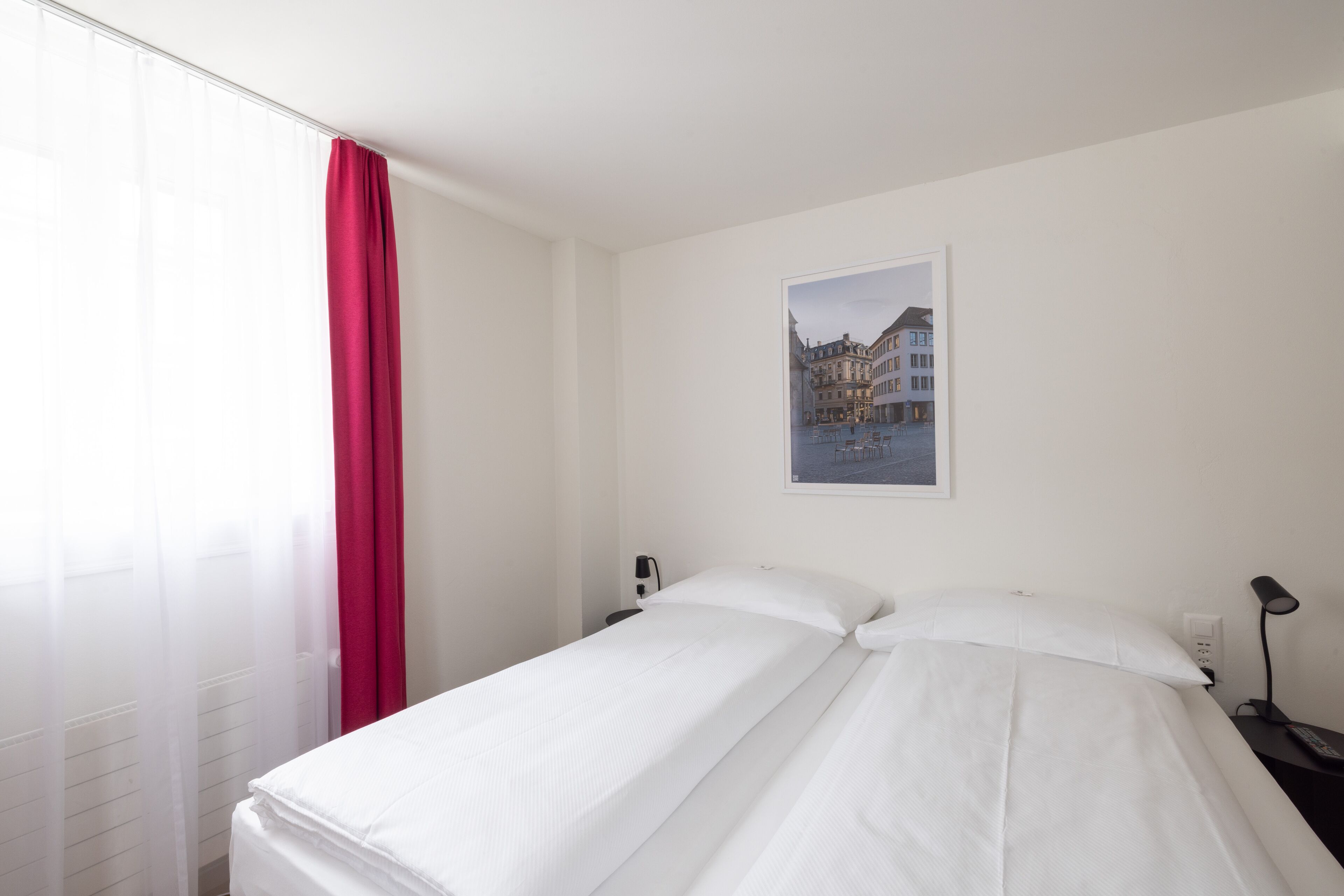 Una habitación de hotel con cama, mesita de noche, lámpara y ventana con cortinas traslúcidas.