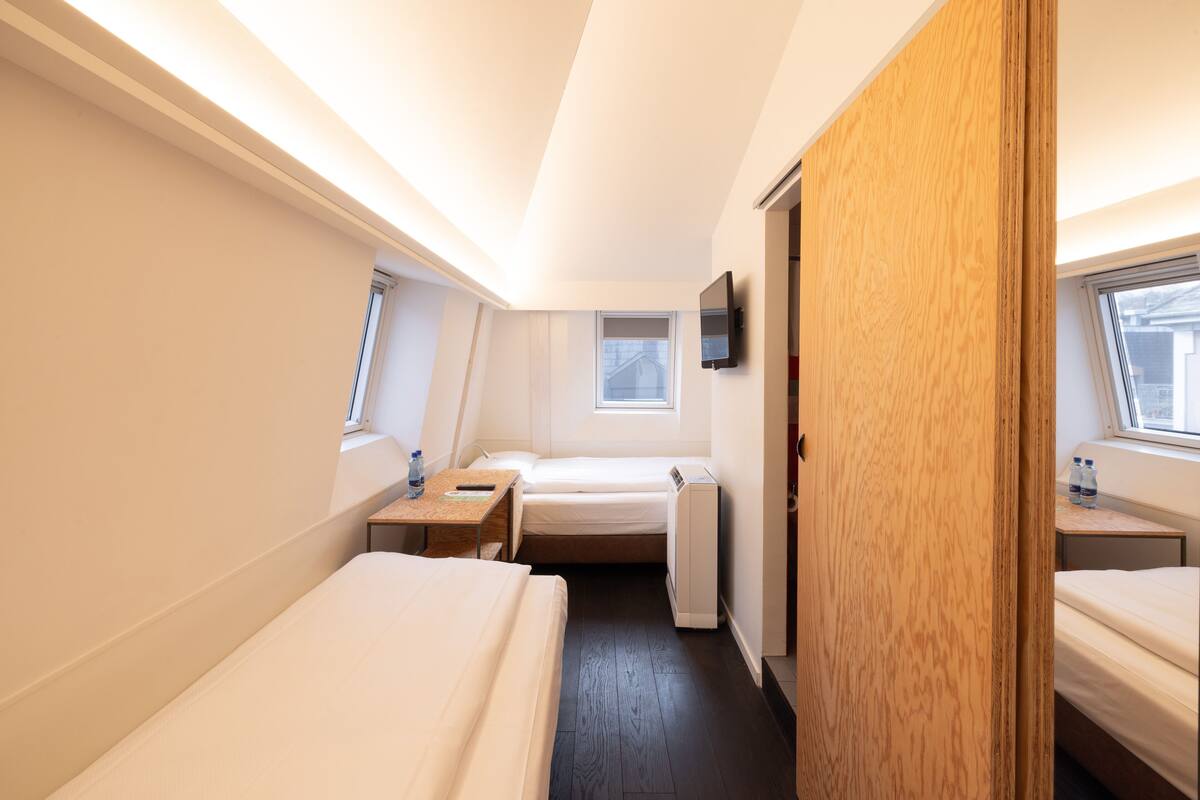 Ein kleines Hotelzimmer mit einem Einzelbett, einem Schreibtisch, einem Stuhl, einem kleinen Tisch, einem Fernseher und einem Fenster.