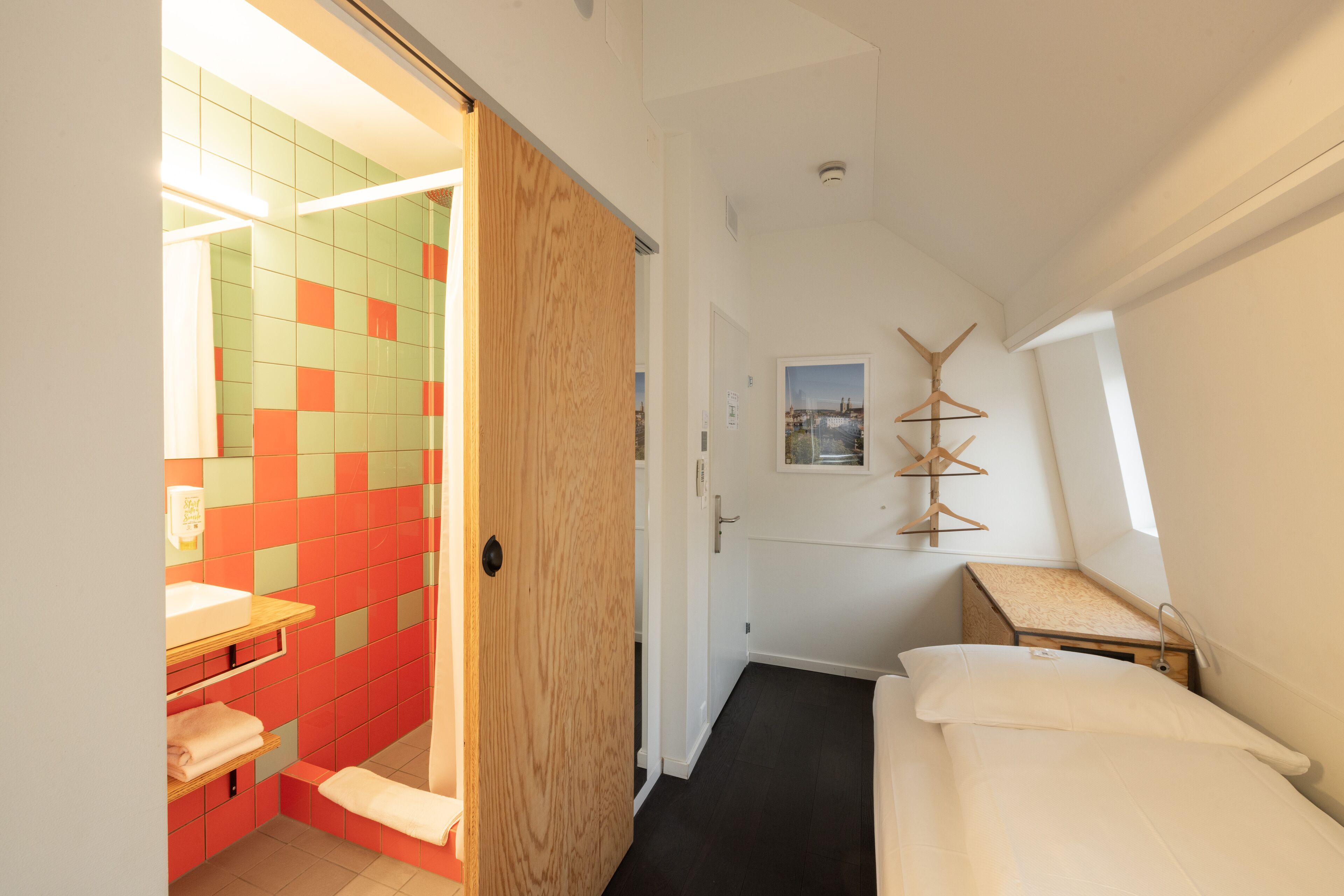A bedroom with a bed, a wooden nightstand, a small table, a window, and a bathroom with a sink and a mirror.